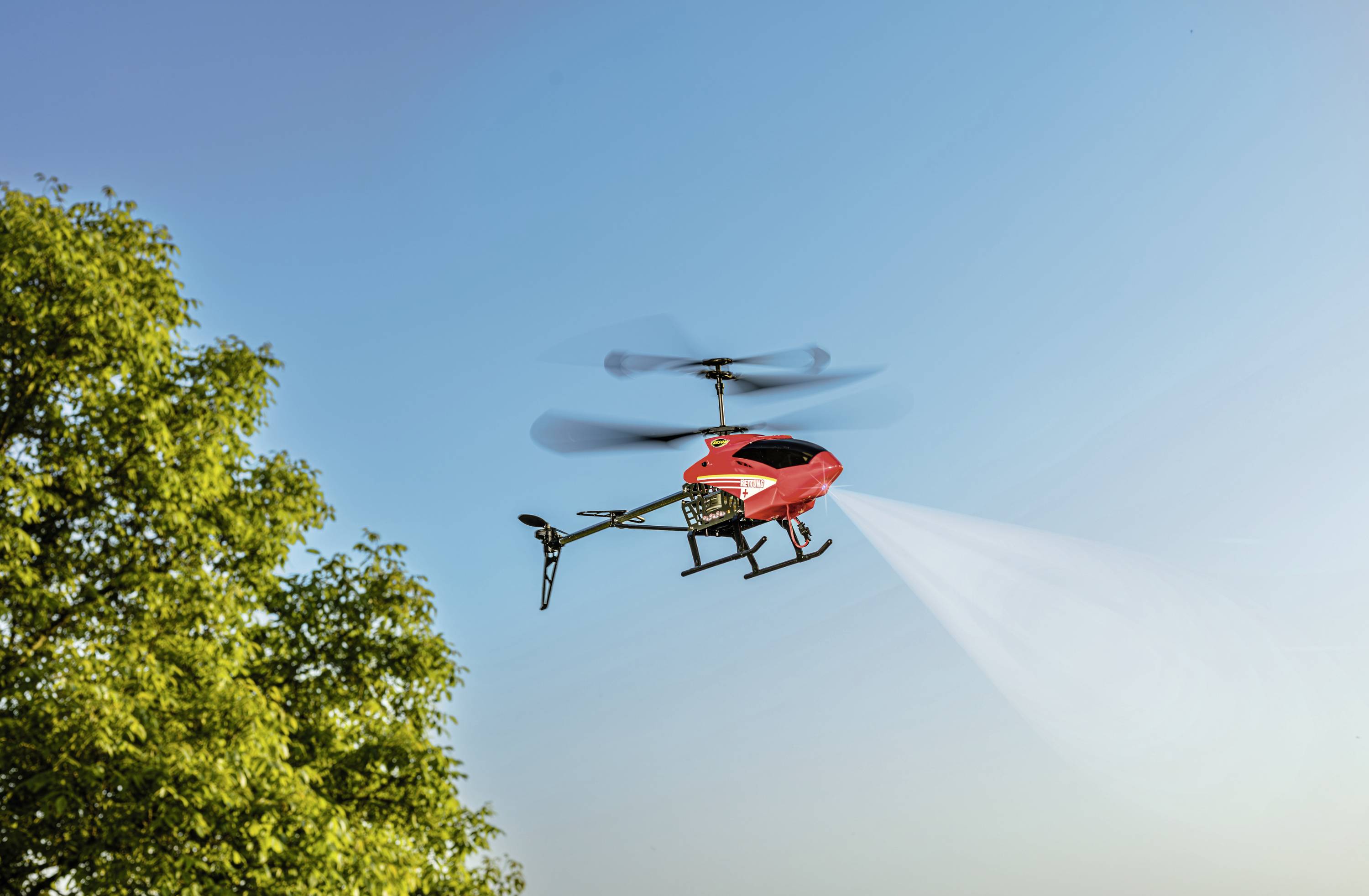 Czerwony dron opryskuje płyn nad polem, w tle błękitne niebo, po lewej stronie drzewo.