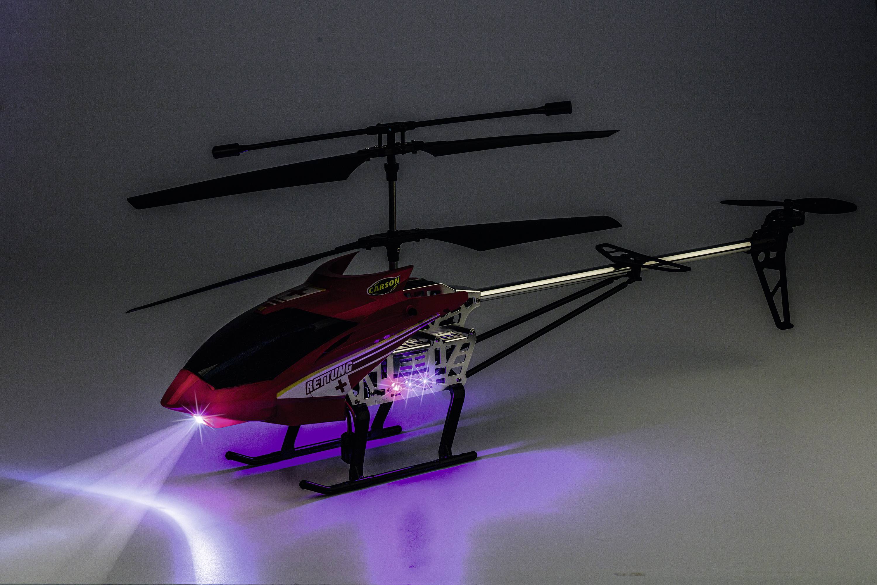 Czerwony zabawkowy helikopter z obracającymi się śmigłami i włączonym przednim reflektorem, stojący na płaskiej powierzchni.