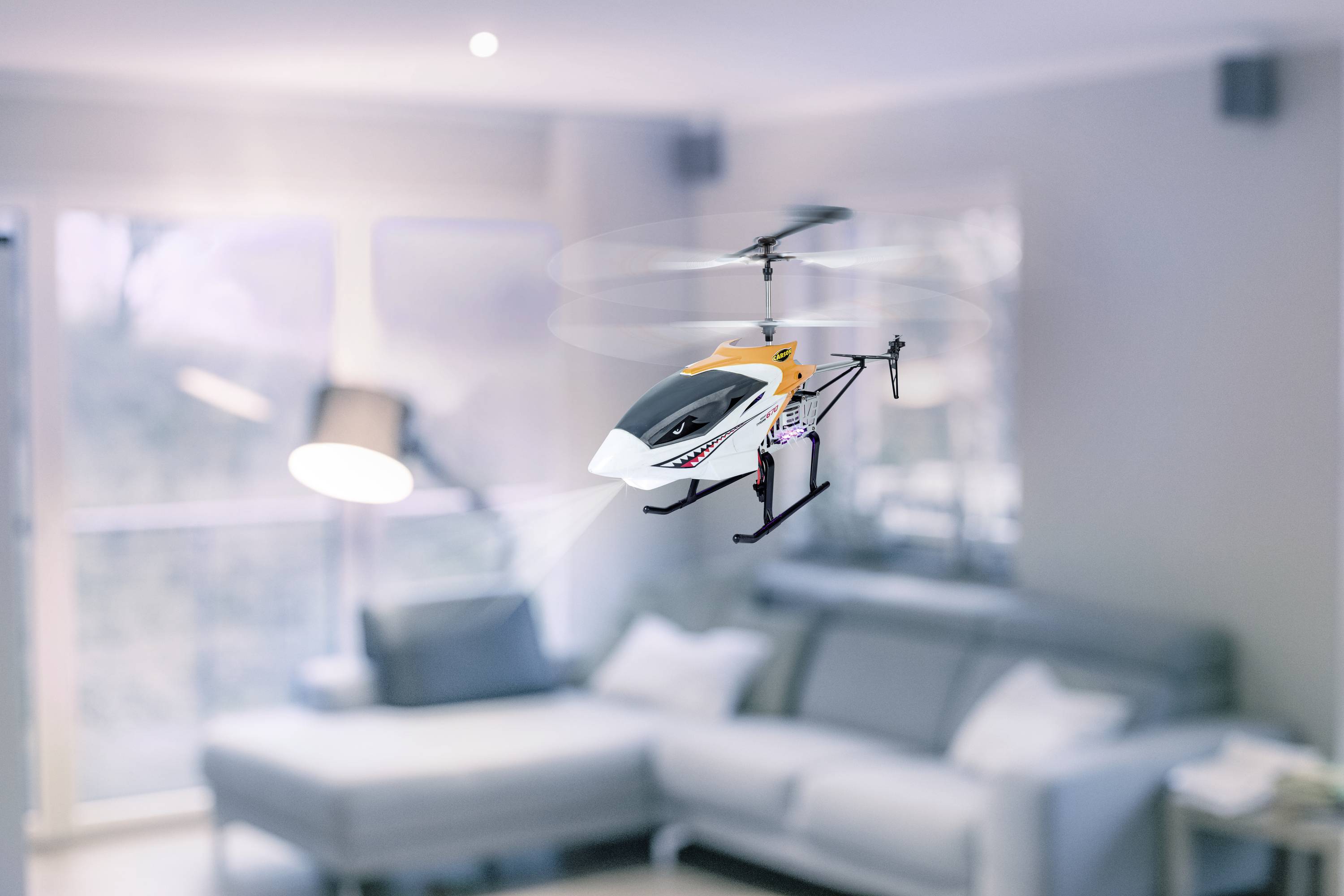 Zdalnie sterowany helikopter lata w nowocześnie urządzonym salonie. W tle widoczna jest kanapa i okna.