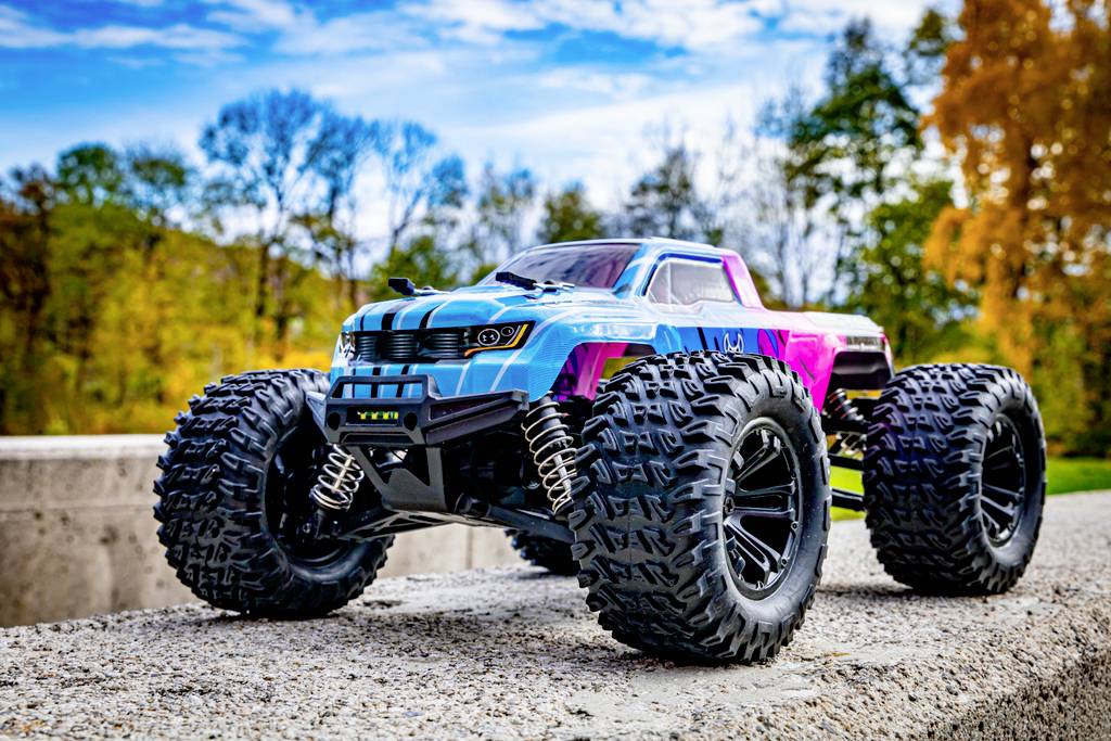 Zdalnie sterowane auto z dużymi oponami stoi na betonowej powierzchni. W tle widać drzewa z jesiennymi liśćmi.