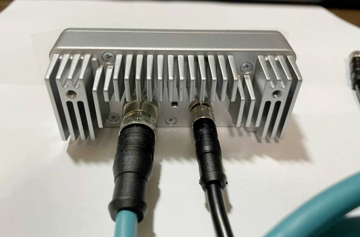 Metalowe urządzenie z żeberkami chłodzącymi i dwoma podłączonymi kablami. Wygląda na urządzenie techniczne, prawdopodobnie wzmacniacz.