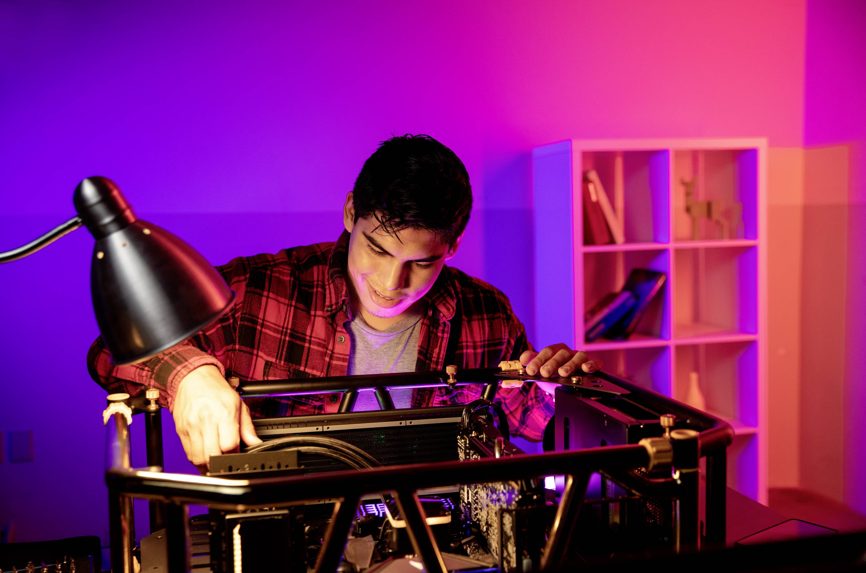 Młody mężczyzna w czerwono-kratowanej koszuli składa komputer, oświetlony kolorowymi światłami w nowoczesnym pomieszczeniu.