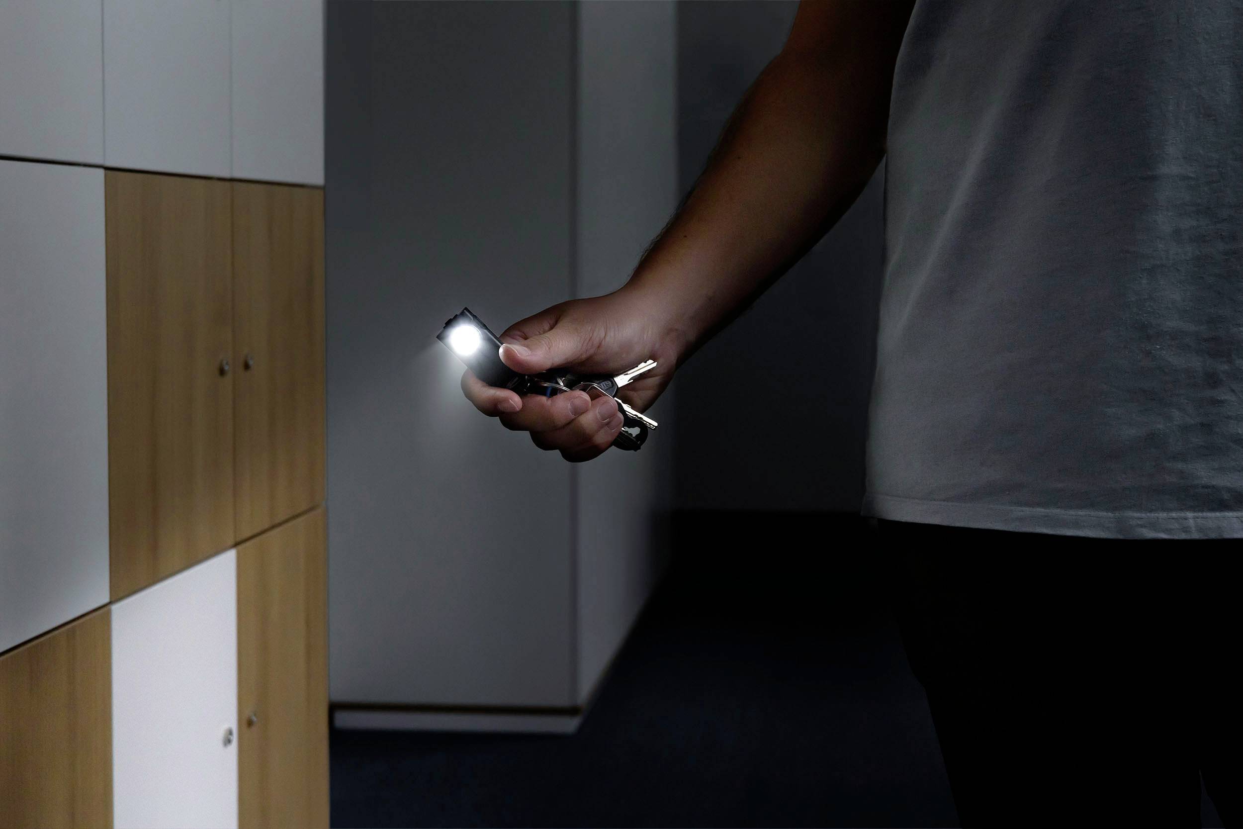 Jedna osoba oświetla ciemny korytarz obok szafy za pomocą małej latarki na breloku.