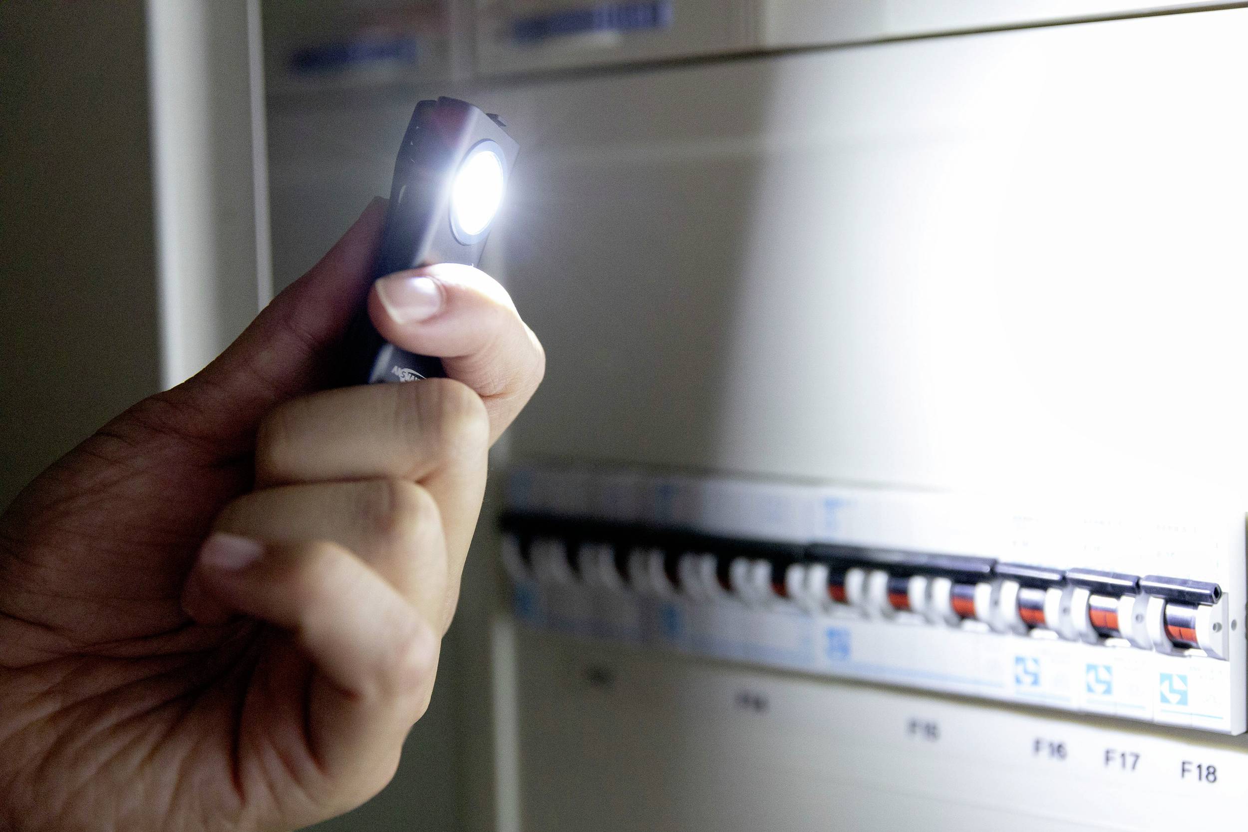 Jedna ręka trzyma latarkę przed otwartą skrzynką bezpieczników z kilkoma bezpiecznikami. Pomieszczenie jest słabo oświetlone.