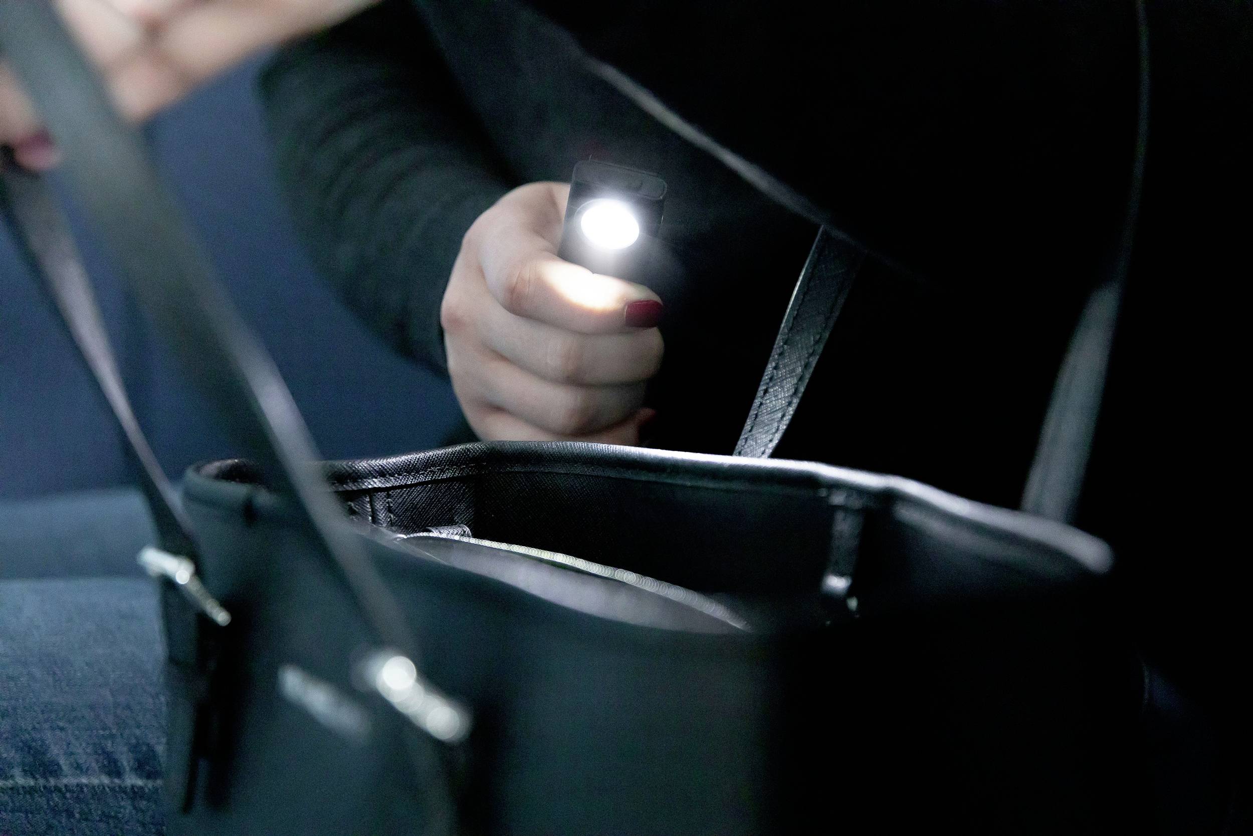 Osoba świeci małą latarką do otwartej czarnej torebki, szukając jakiegoś przedmiotu.