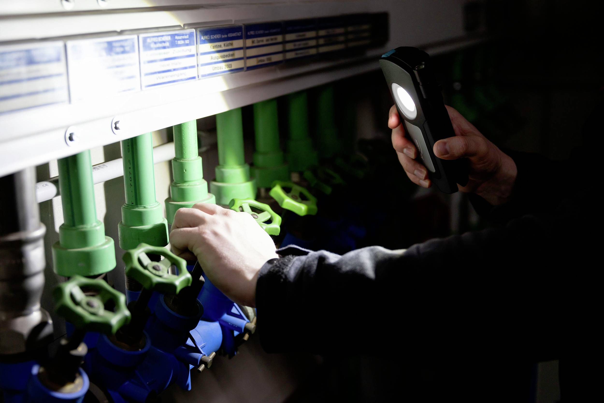 Ręka trzyma latarkę i sprawdza kilka zaworów oznaczonych na niebiesko i zielono, znajdujących się na rurociągu.