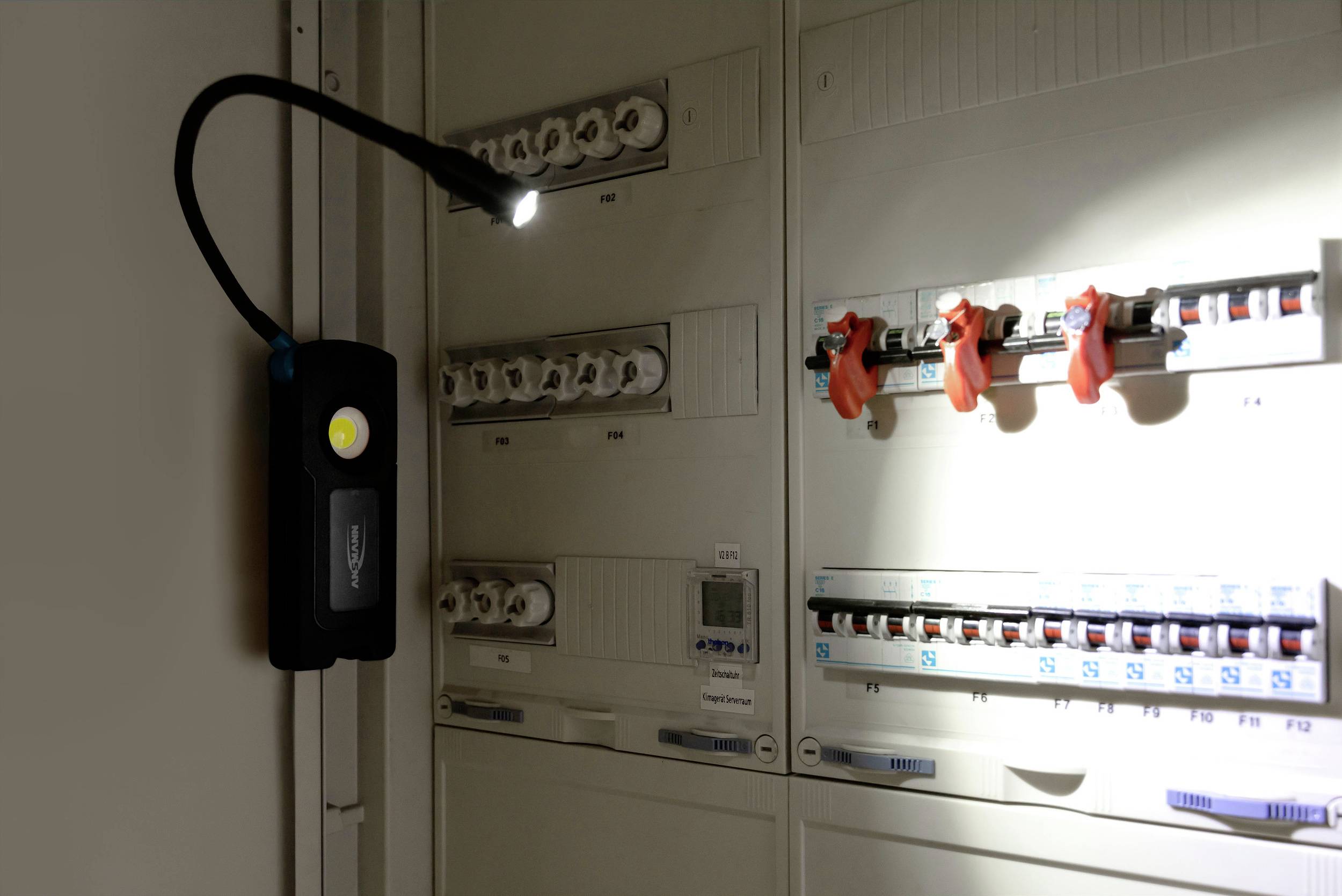 W ciemnym pomieszczeniu przenośna lampa rzuca światło na elektryczną szafkę rozdzielczą, ukazując bezpieczniki i przełączniki.