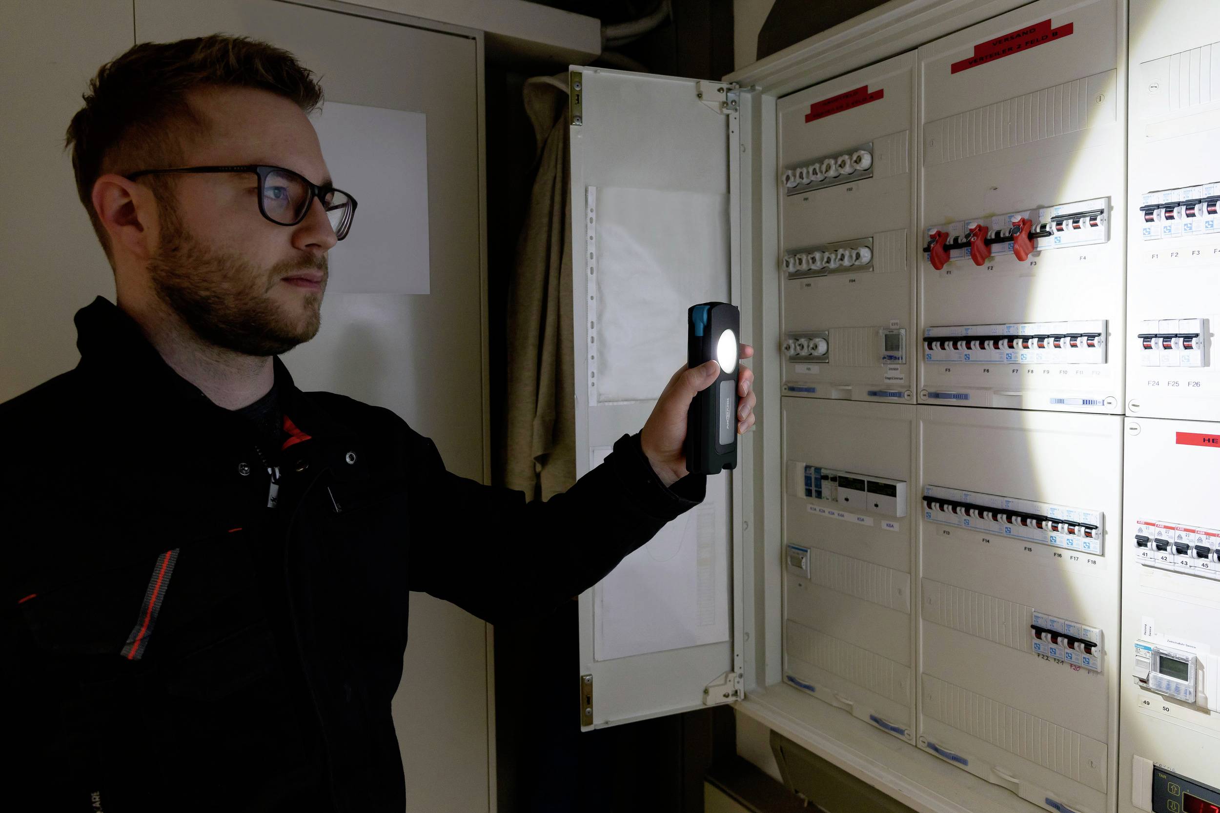 Mężczyzna sprawdza otwarty rozdzielnik elektryczny za pomocą miernika kieszonkowego. Ma na sobie okulary i ciemne ubranie.