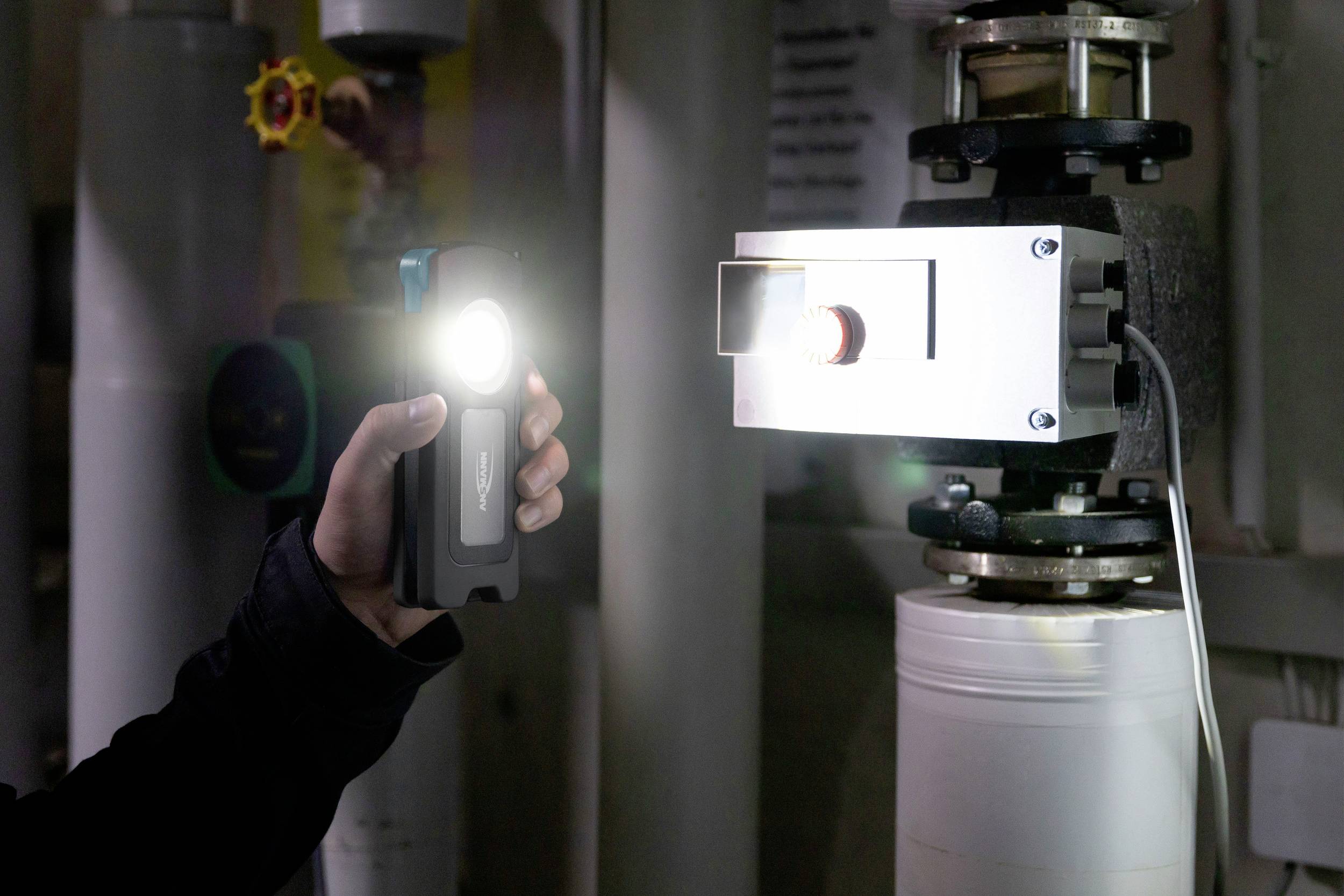 Jedna osoba trzyma świecącą latarkę przy instalacji technicznej z zaworami i rurami. Otoczenie jest słabo oświetlone.