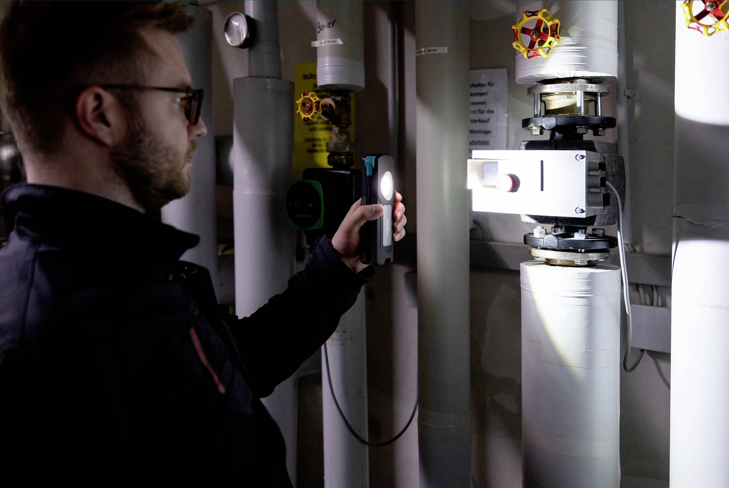 Osoba sprawdza techniczne przewody rurowe latarką w ciemnym pomieszczeniu. Widocznych jest kilka rur i zaworów.