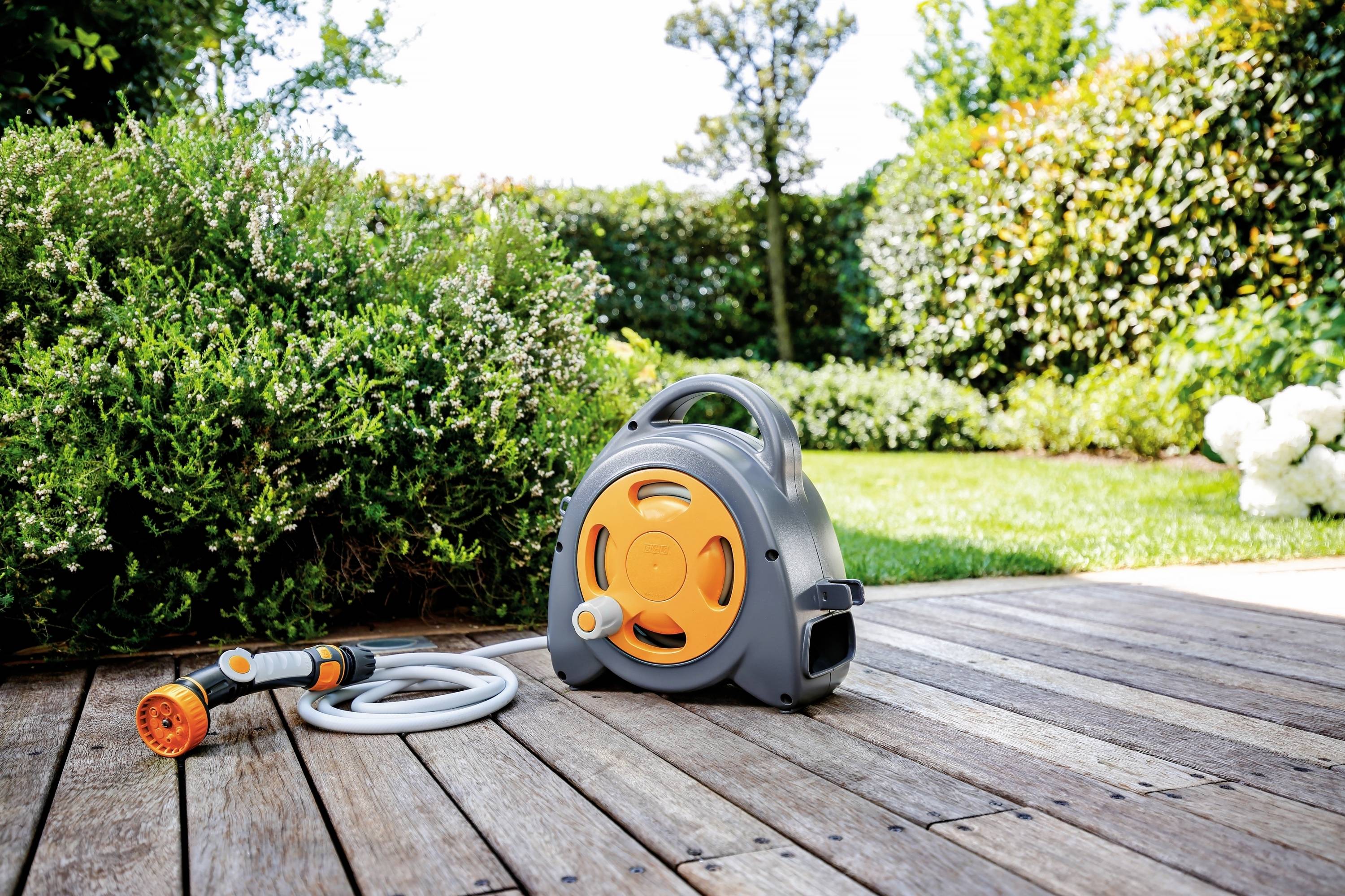 Wąż ogrodowy zwinięty w uchwycie na drewnianym tarasie z zielonym tłem ogrodu.