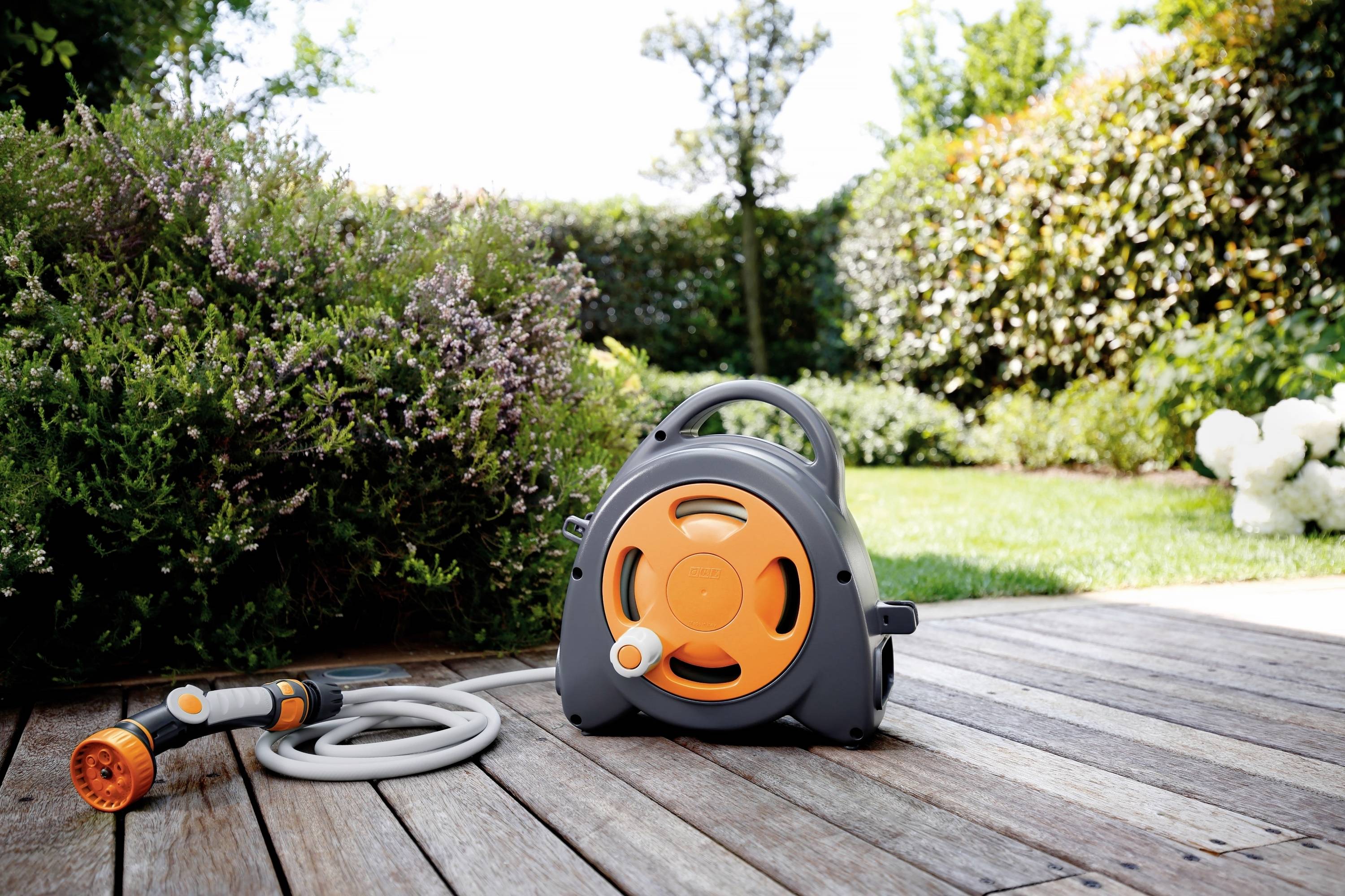 Wąż ogrodowy na stojaku nawijającym, umieszczony na drewnianym tarasie, otoczony zielenią i żywopłotem w tle.