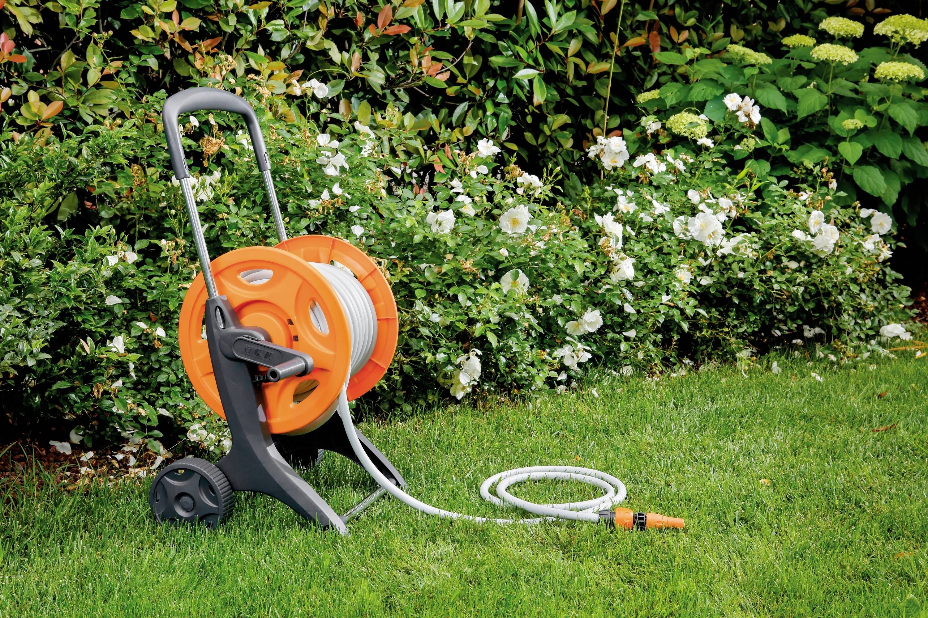 Pomarańczowy wąż ogrodowy na wózku stoi na zielonym trawniku przed rabatką z kwitnącymi roślinami.