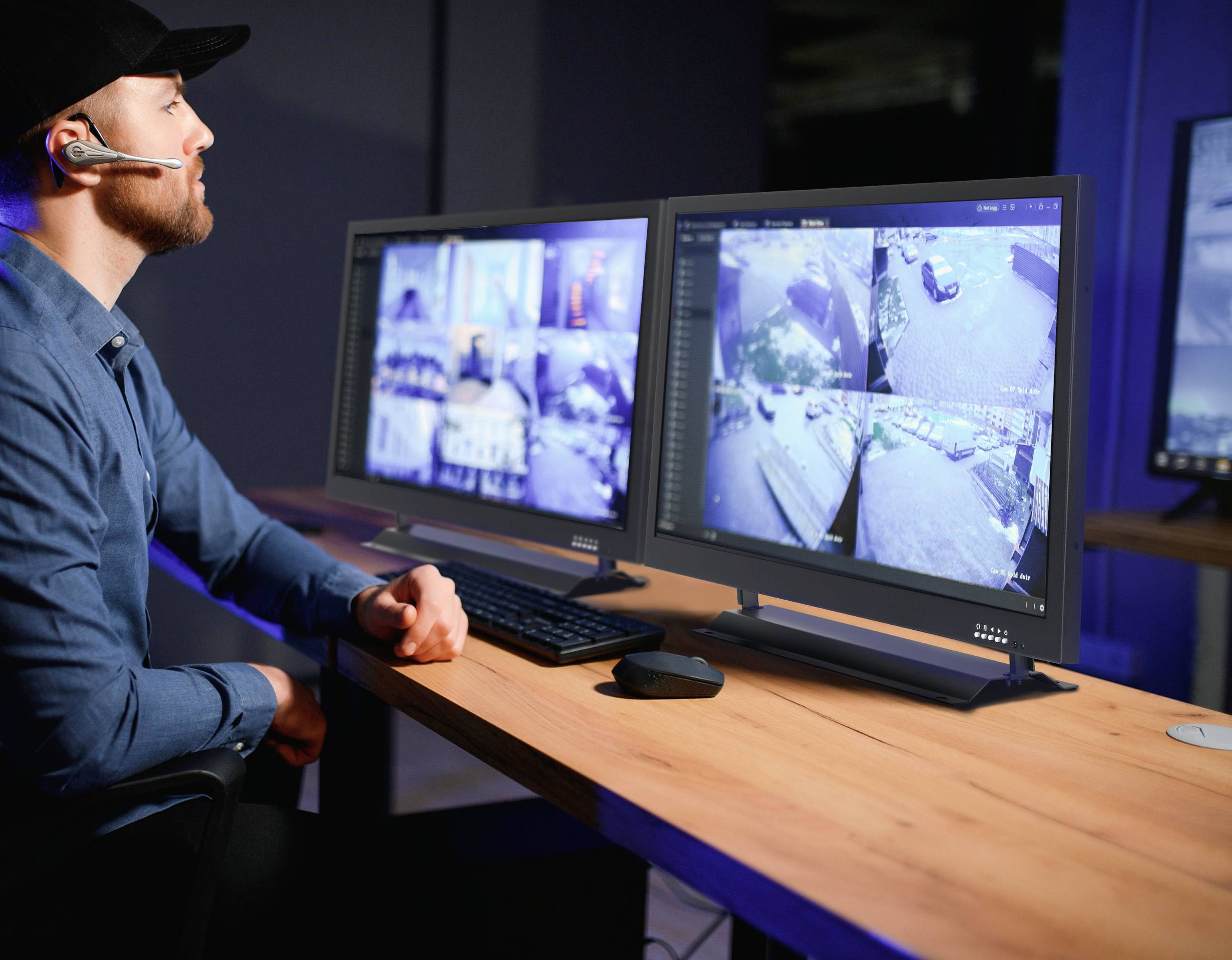 Mężczyzna w zestawie słuchawkowym siedzi przed dwoma monitorami wyświetlającymi obrazy z kamer monitoringu w pomieszczeniu kontrolnym.
