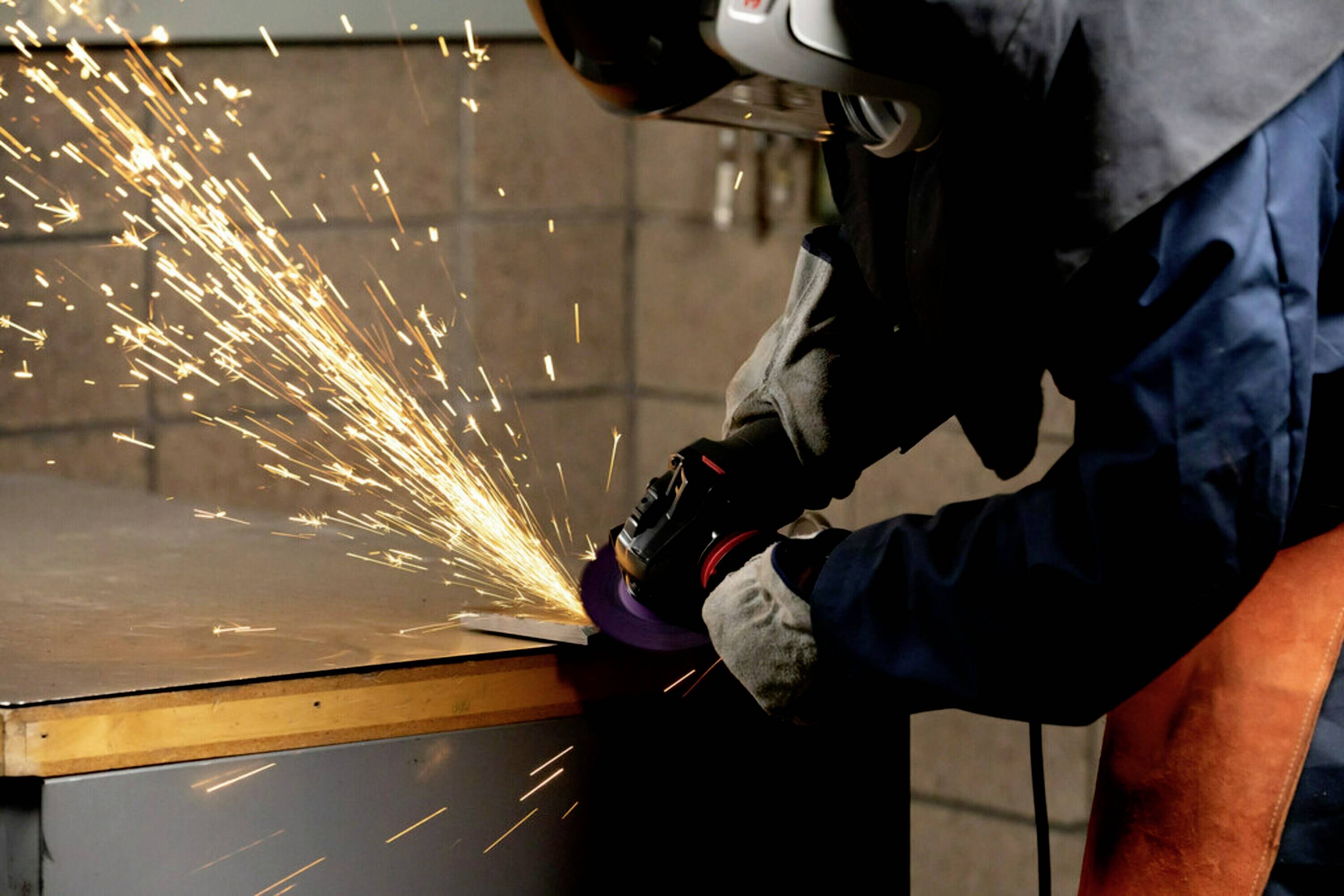 Osoba szlifuje metal przy stole, iskry się sypią. Ma na sobie odzież ochronną i kask.