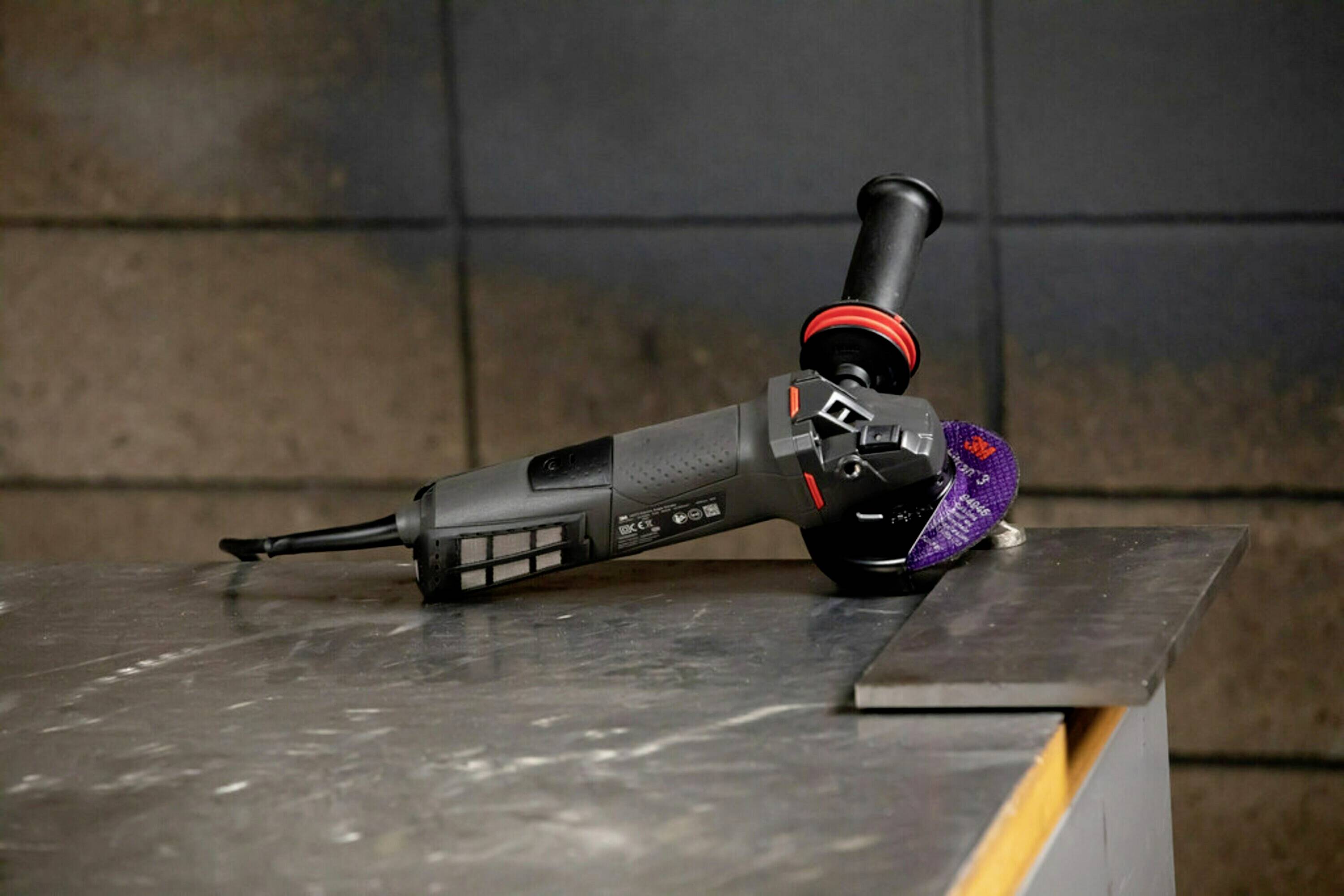 Szlifierka kątowa leży na metalowym stole. Posiada ergonomiczny uchwyt, a tarcza szlifierska dotyka metalowej płyty.