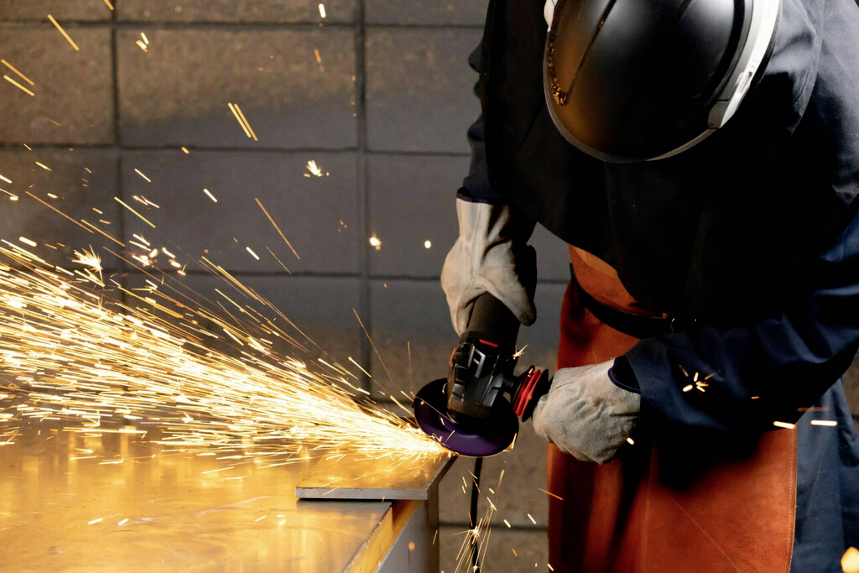 Osoba szlifuje metal szlifierką kątową, a wokół lecą iskry. Ma na sobie odzież ochronną, w tym kask i rękawice.