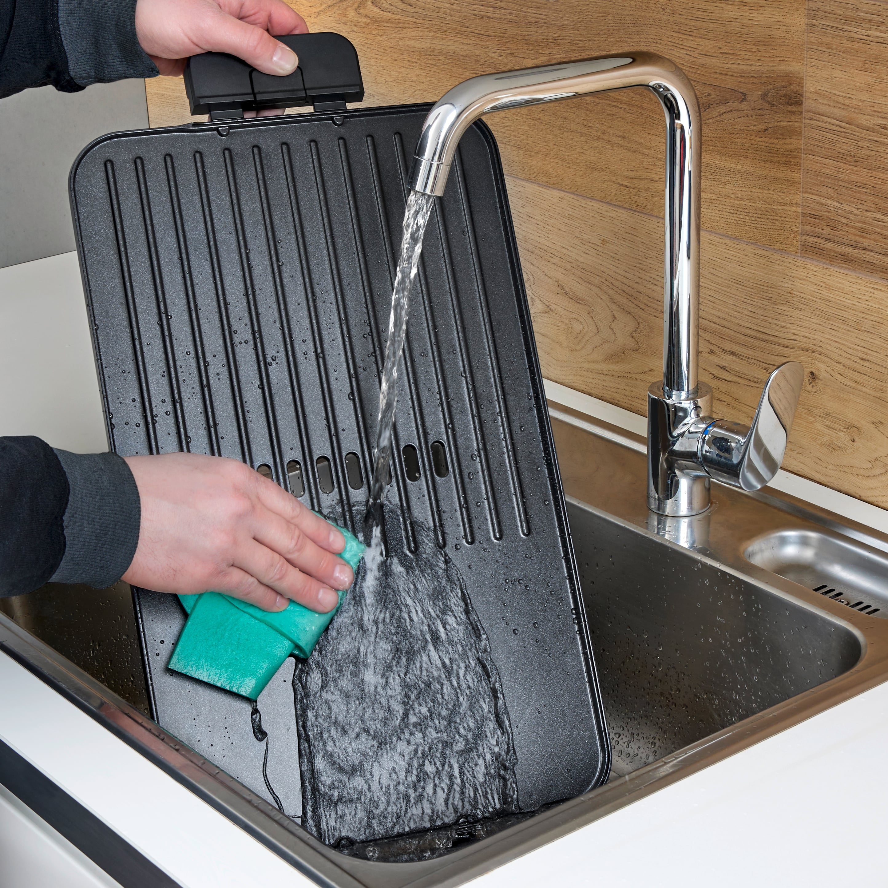 Osoba czyści żelazko do gofrów zielonią gąbką pod bieżącą wodą w zlewie.