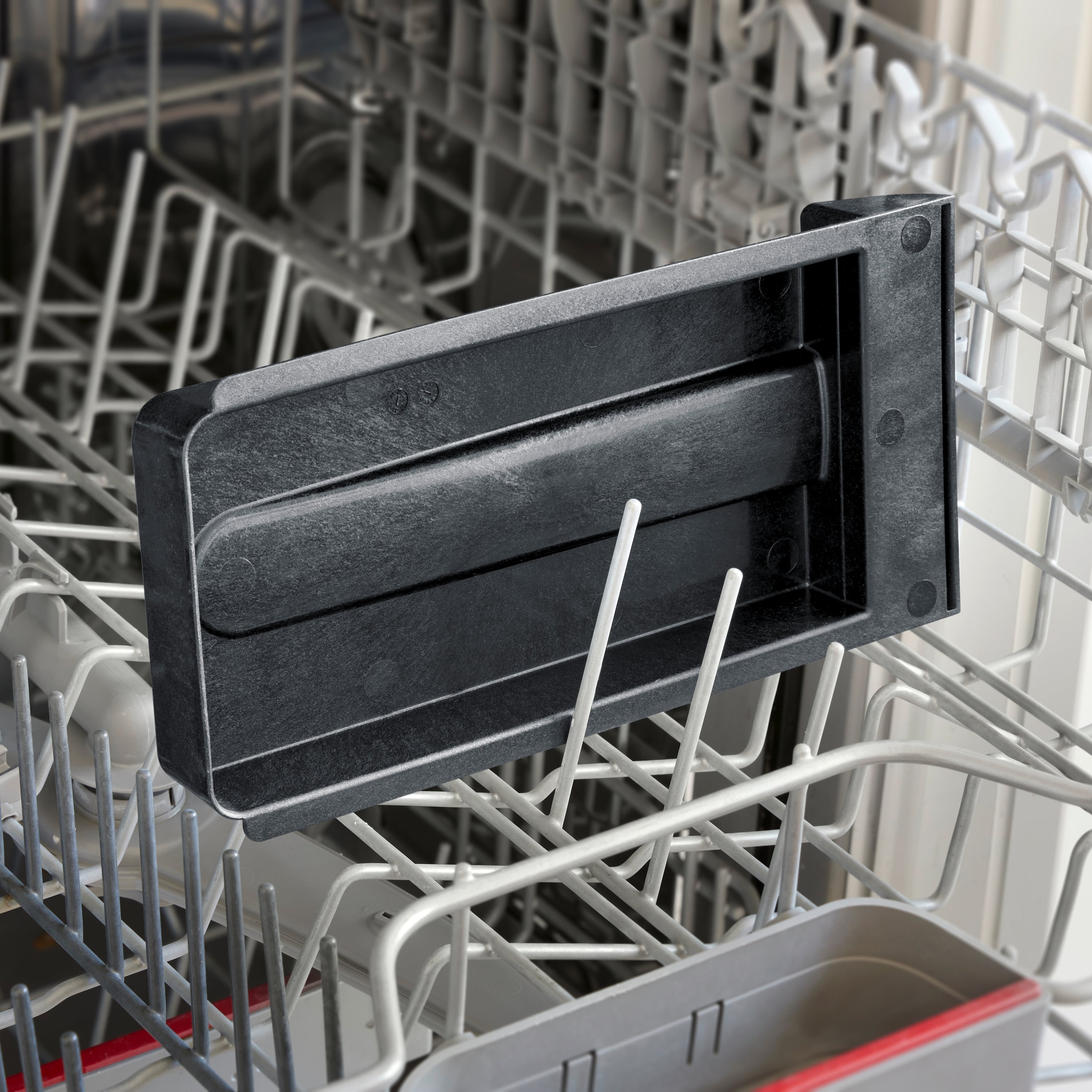 Czarny obiekt z tworzywa sztucznego umieszczony w dolnej części otwartej zmywarki. Charakteryzuje go proste wzornictwo.