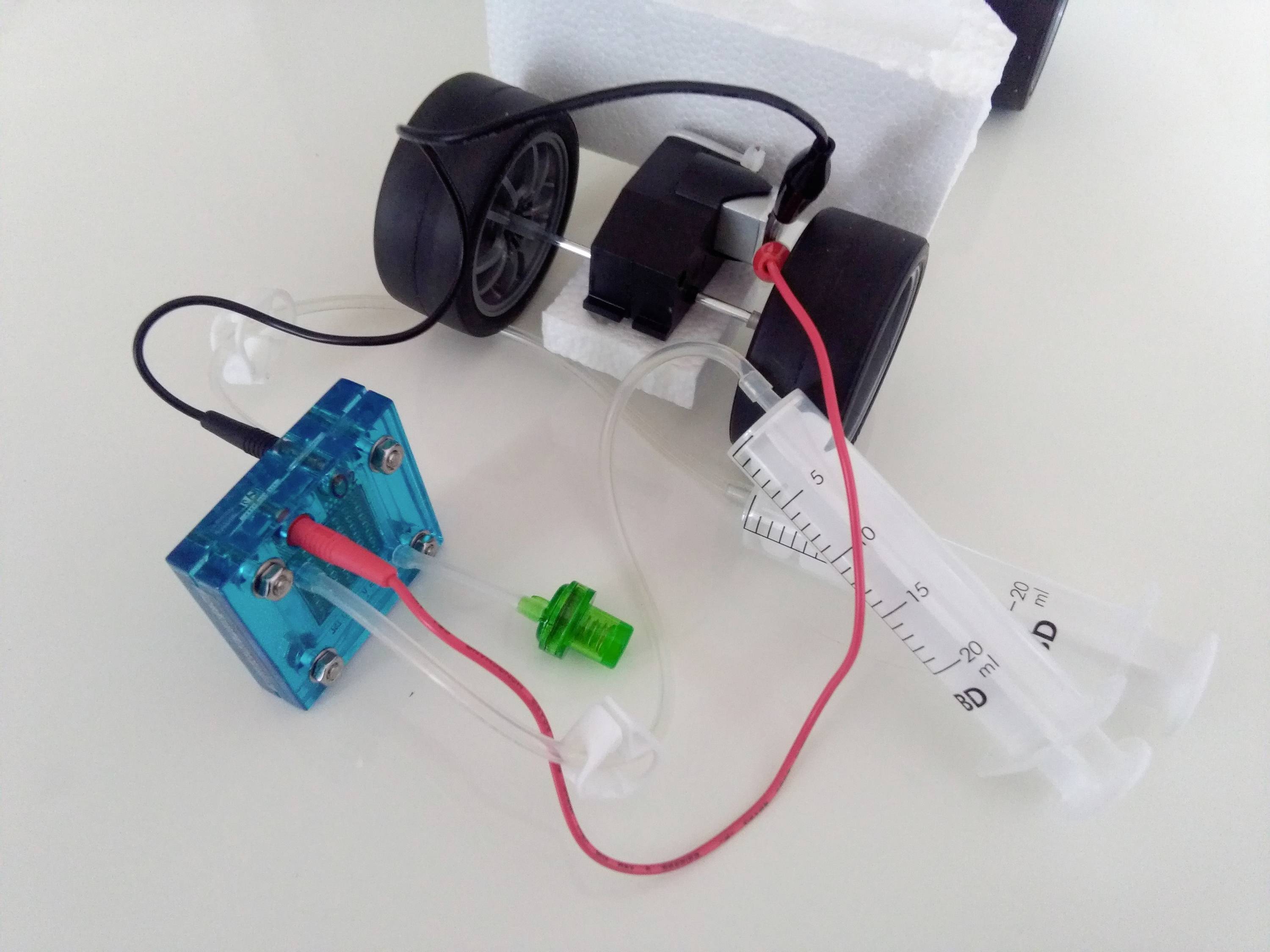 Mały pojazd z dwoma kołami, połączony ze strzykawką i modułem elektrycznym, prezentuje prosty układ hydrauliczny.