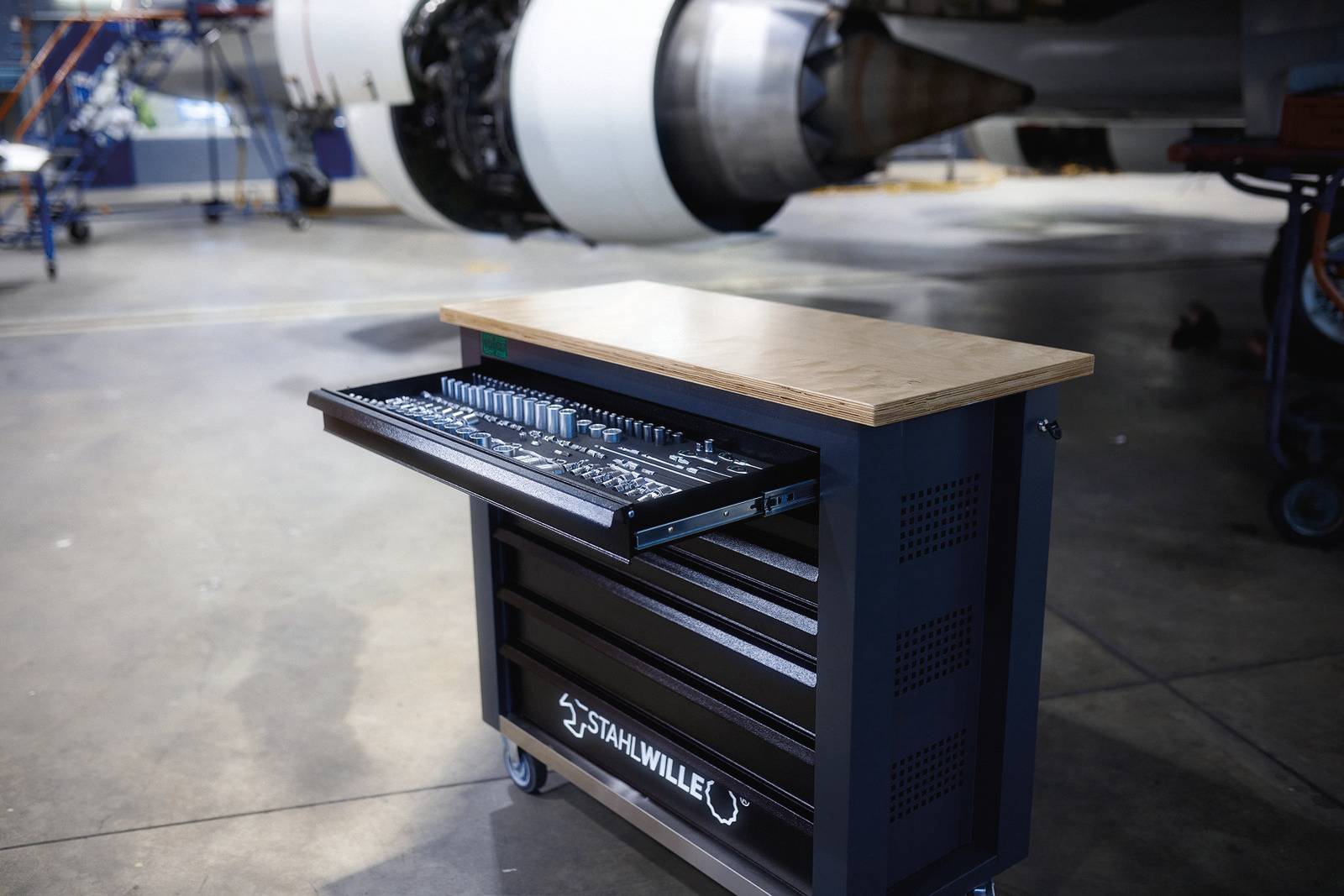 Wózek narzędziowy z otwartą szufladą przed silnikiem lotniczym w hali warsztatowej, prezentujący różnorodne narzędzia do obsługi technicznej samolotów.