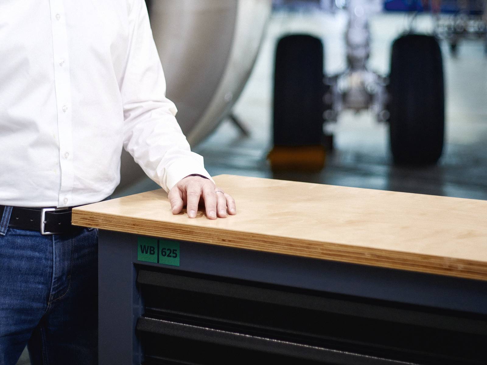 Osoba w białej koszuli stoi obok stołu warsztatowego w warsztacie. W tle niewyraźnie widoczne są koła samolotowe.