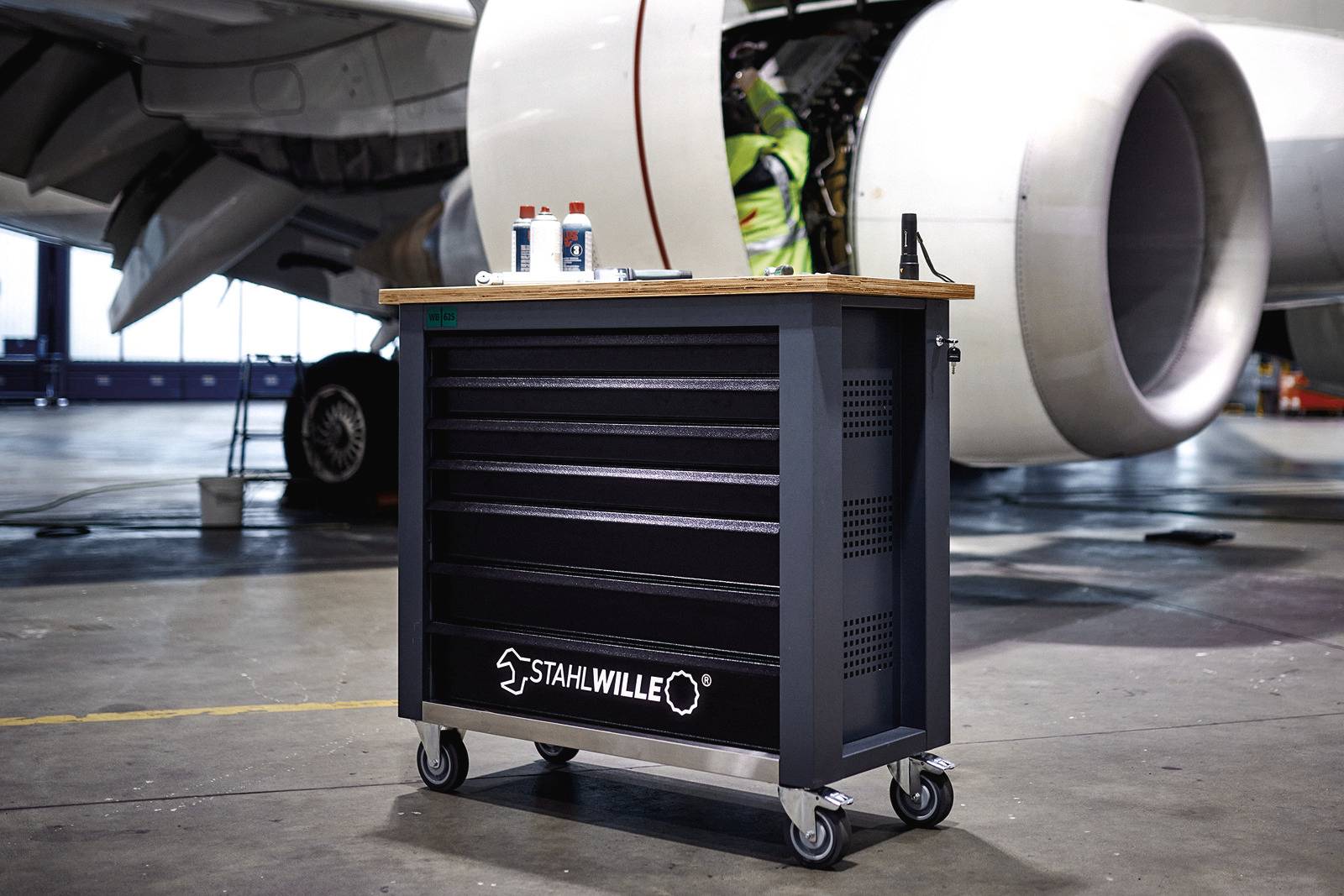 Wózek narzędziowy firmy Stahlwille stoi w hangarze przed silnikiem lotniczym. Technik pracuje przy silniku.