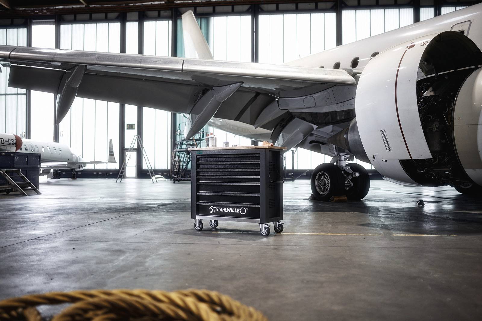 Wóz serwisowy stoi w hangarze przed samolotem. Samolot ma otwarte klapy, co wskazuje na trwające prace konserwacyjne.