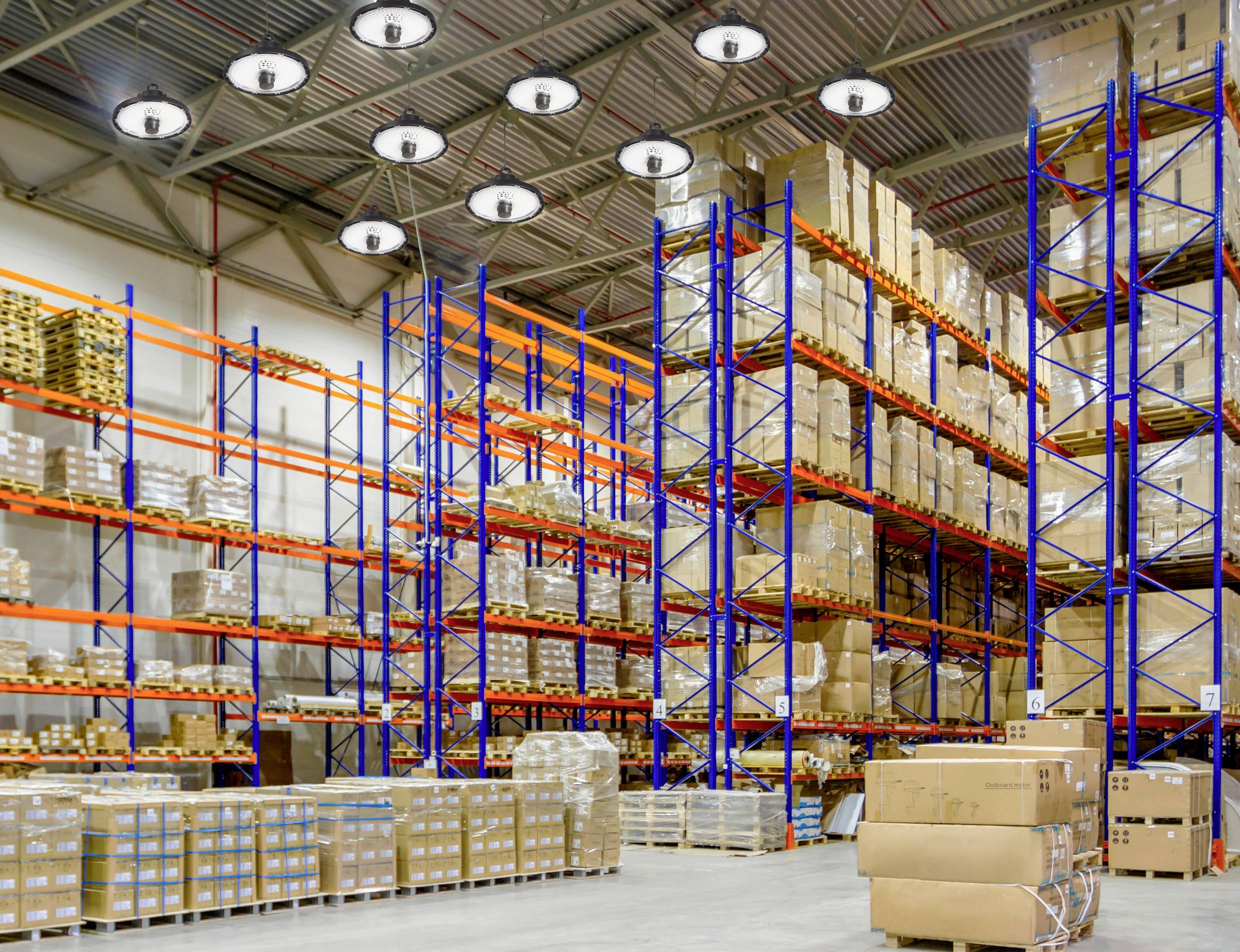 Duży magazyn z wysokimi regałami, wypełniony skrzynkami i paczkami. Jasne oświetlenie przemysłowe i palety na podłodze.