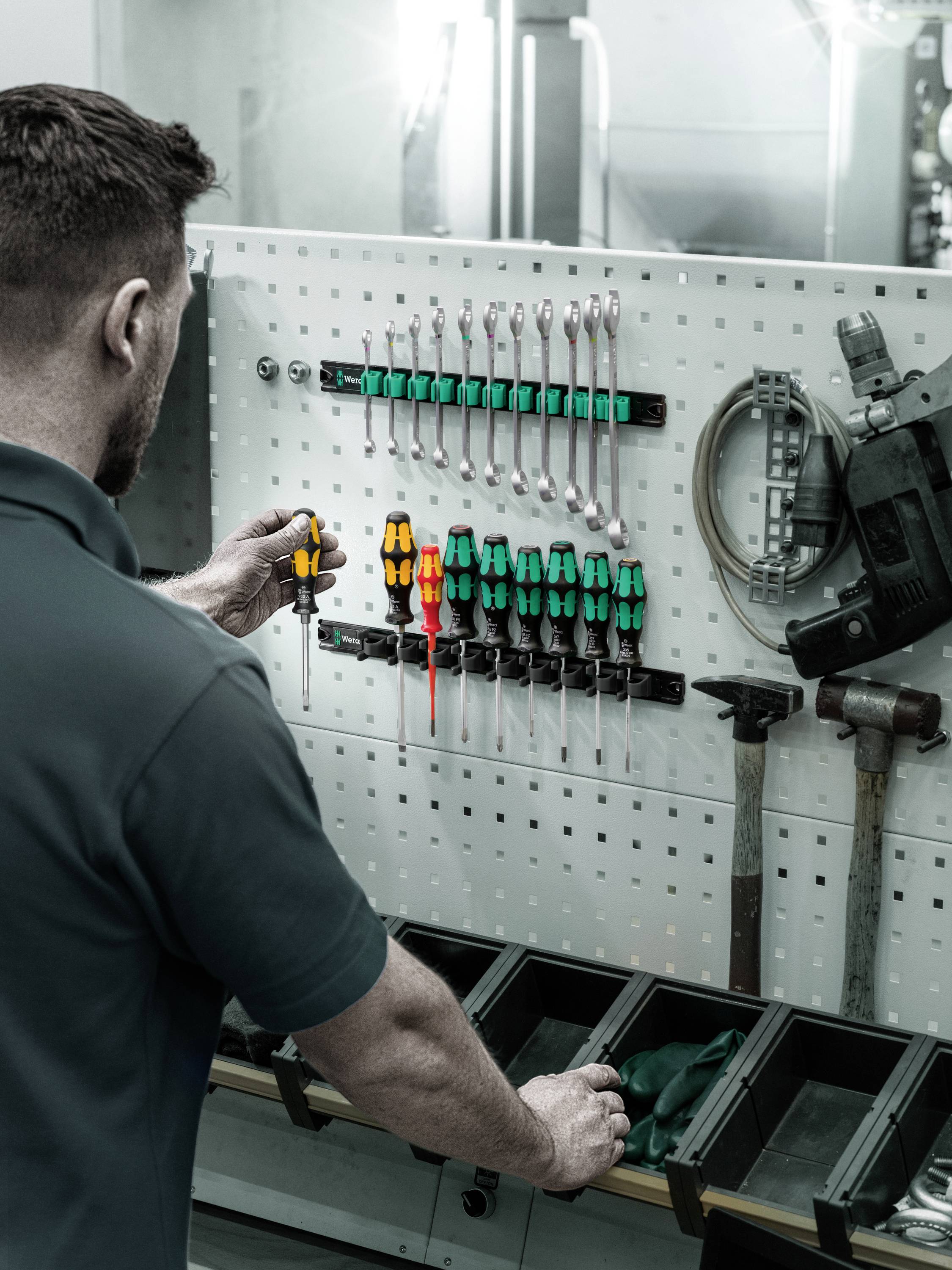 Mężczyzna rozmieszcza narzędzia na tablicy z otworami, w tym śrubokręty, klucze i elektronarzędzie, w warsztacie.