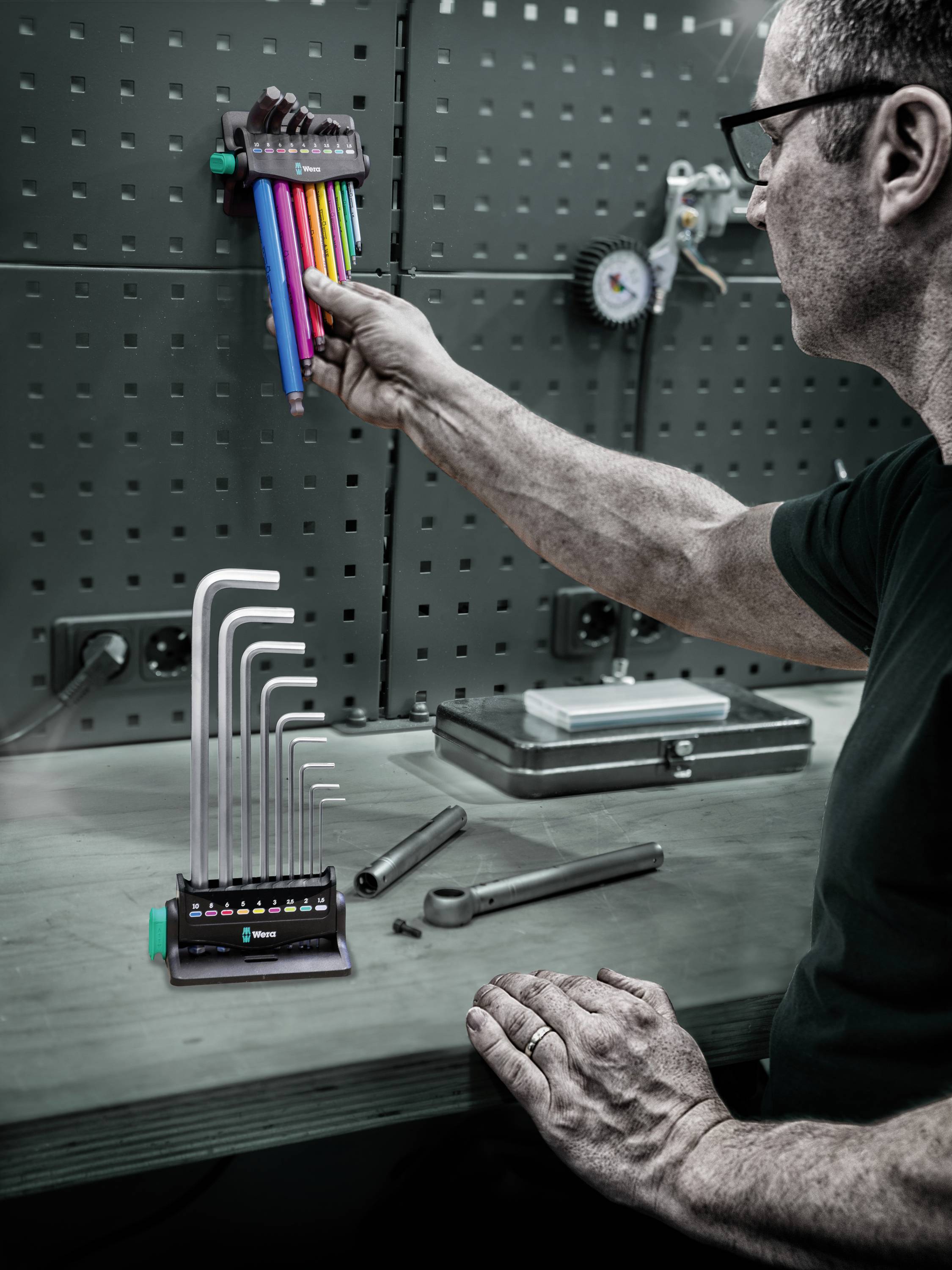 Mężczyzna rozmieszcza kolorowe klucze imbusowe na ścianie warsztatowej i kładzie rękę na dwóch kluczykach leżących na stole.