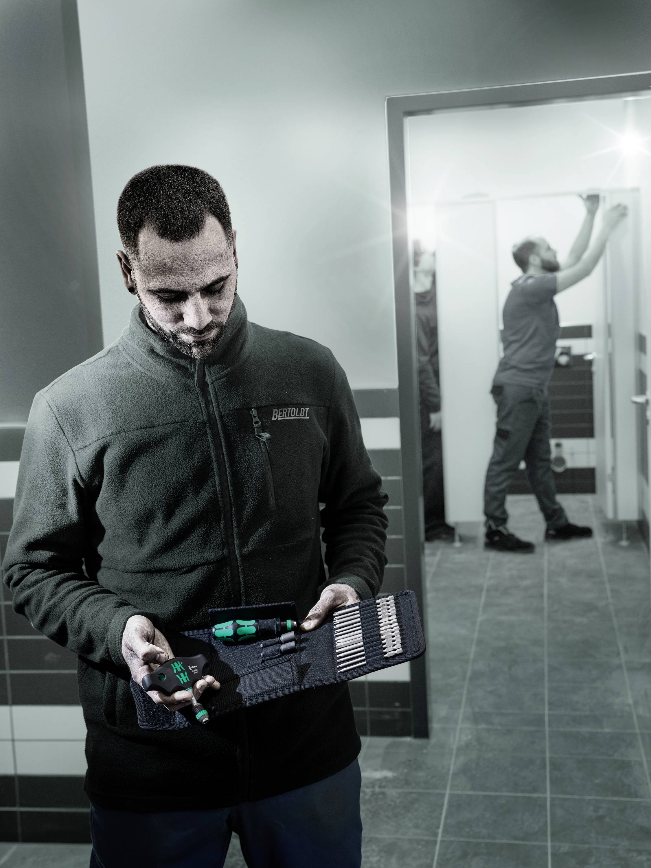 Mężczyzna trzyma walizkę z narzędziami, podczas gdy drugi mężczyzna w tle pracuje przy suficie. Znajdują się w jasnym pomieszczeniu.