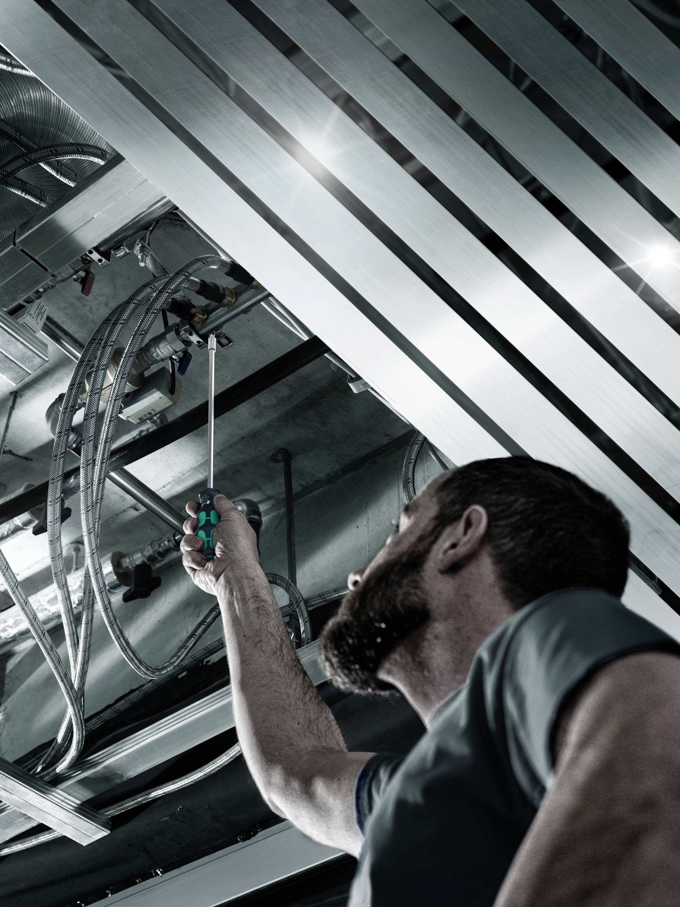 Mężczyzna pracuje przy suficie instalacji przemysłowej, trzymając narzędzie i sprawdzając rury oraz kable w środowisku technicznym.