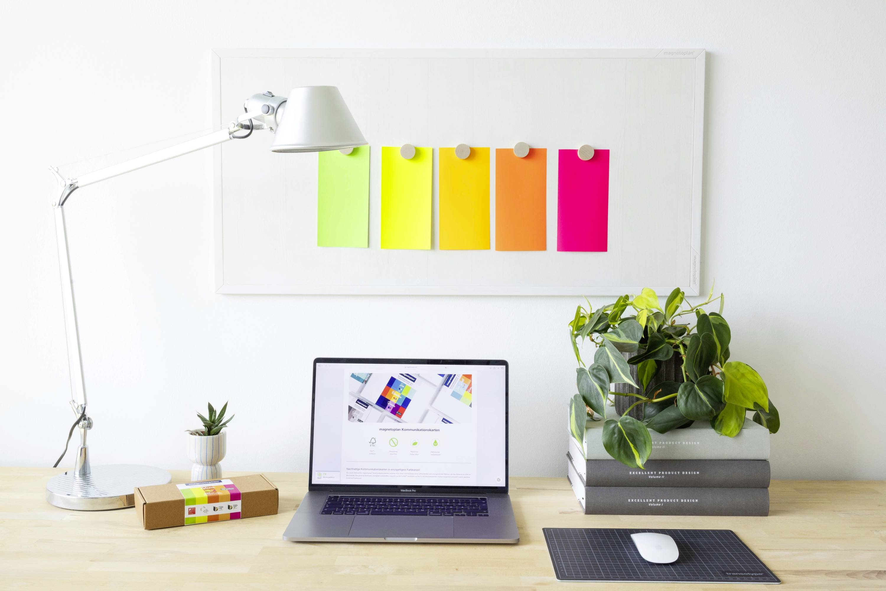 Biały biurko z otwartym laptopem, zieloną rośliną, małym kaktusem, lampką i kolorowymi kartkami przypiętymi do tablicy.