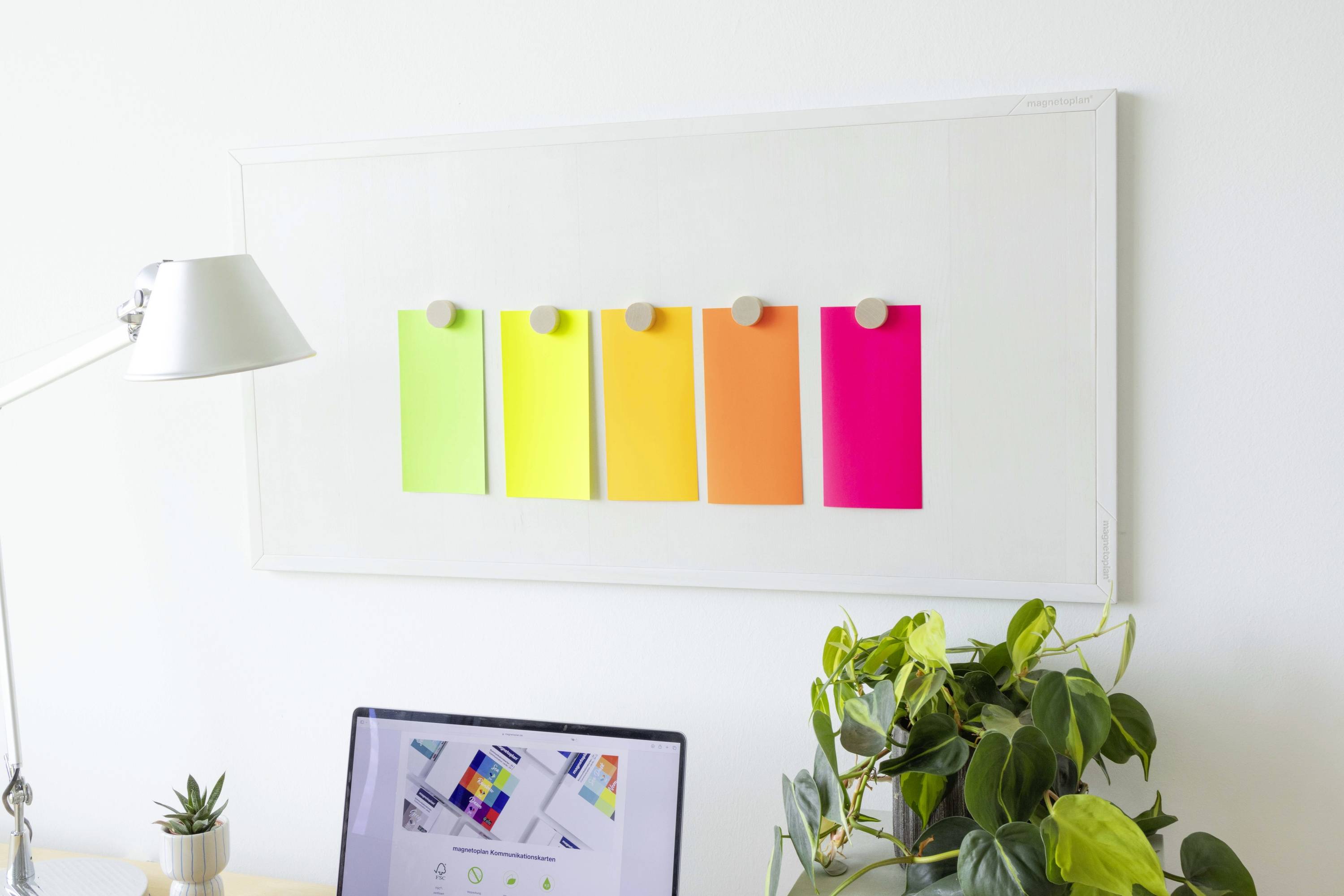 Biurko z lampą, laptopem i roślinami na pierwszym planie; w tle wisi tablica z kolorowymi karteczkami samoprzylepnymi.