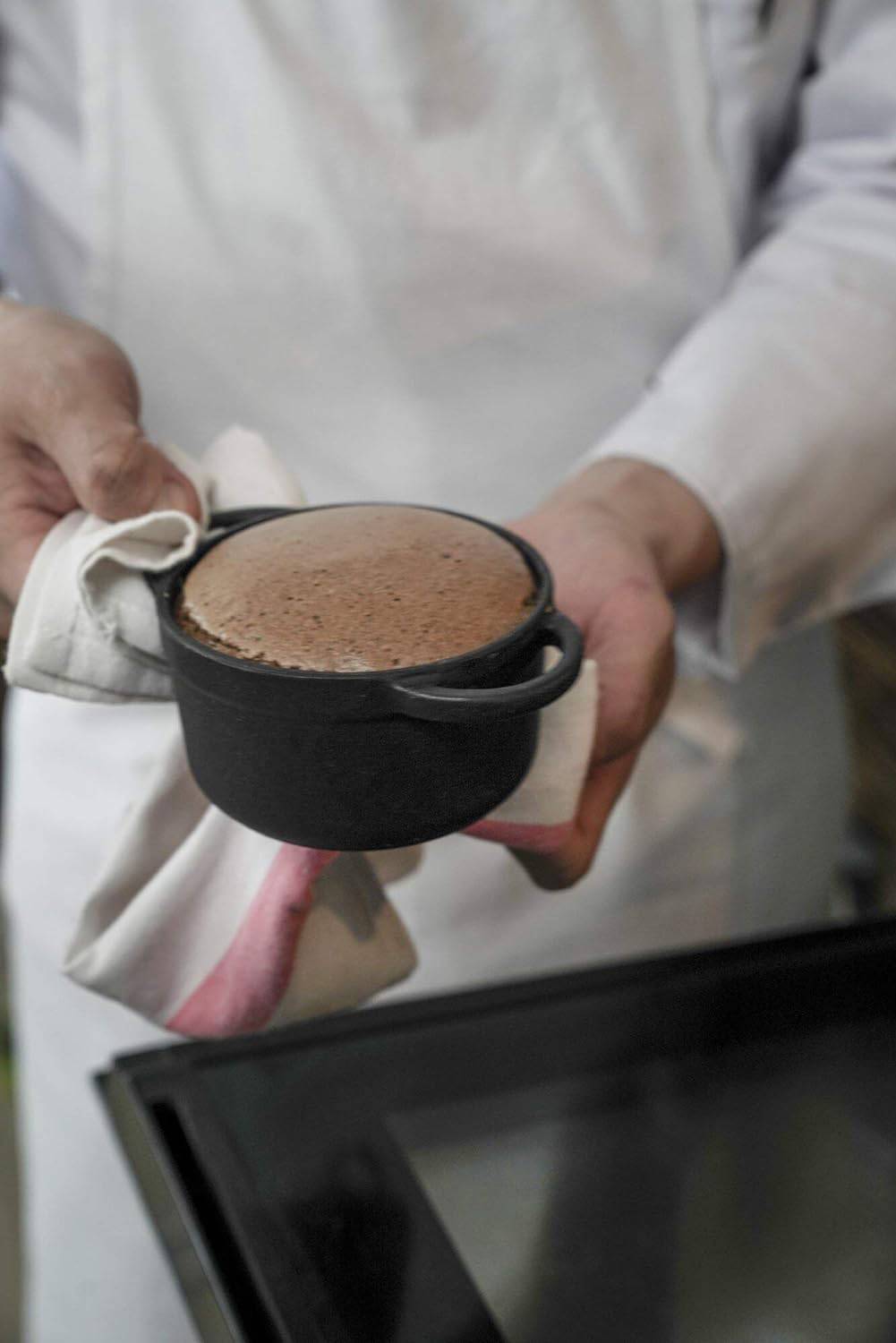 Osoba ubrana na biało trzyma ciepłą, świeżo upieczoną czekoladową blachę w czarnej formie do pieczenia, przykrytą tkaniną.