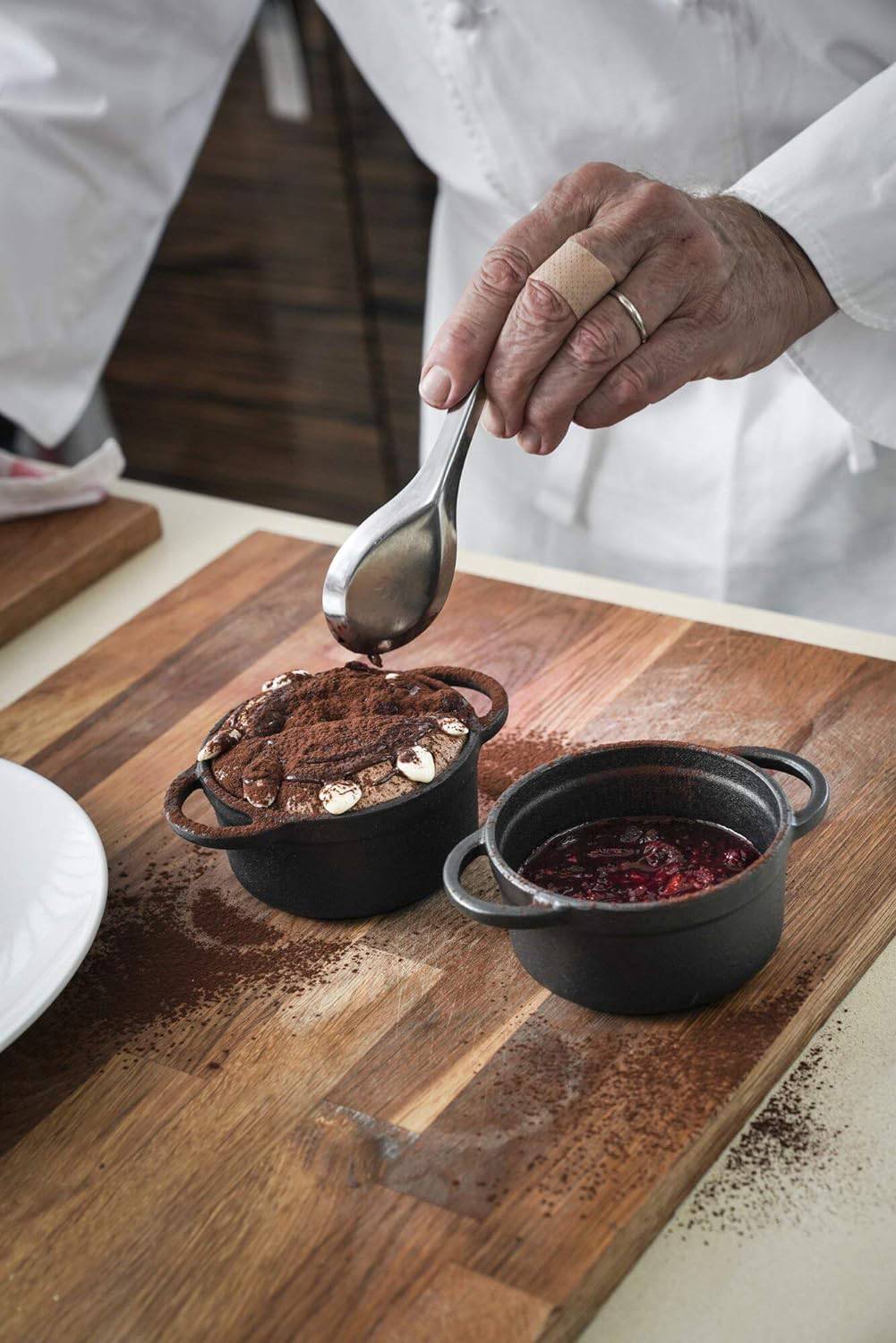 Kucharz posypuje deser czekoladowy kakao, obok małej miseczki z czerwonym sosem na desce drewnianej.