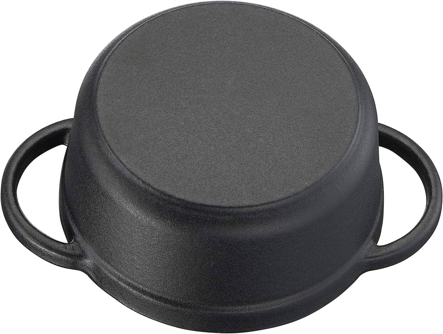 Odwrócony czarny żeliwny garnek z dwoma uchwytami po bokach, leżący na białej powierzchni.