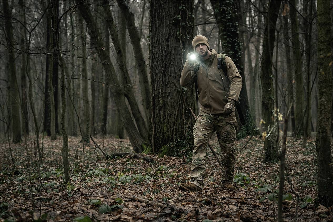 Mężczyzna idzie w nocy przez zalesiony teren, trzymając latarkę, która rozświetla drogę przed nim.