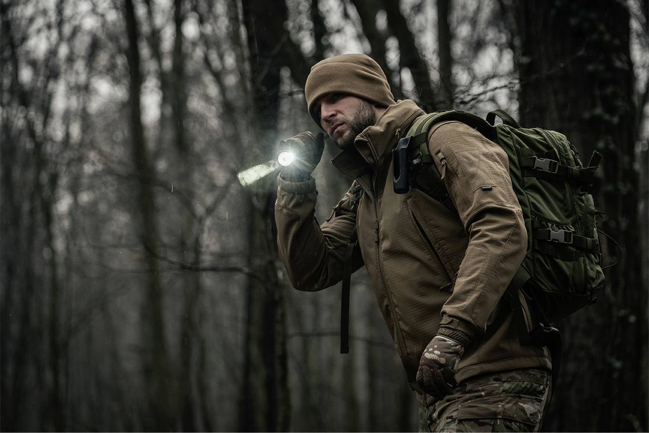 Osoba ubrana w odzież outdoorową i czapkę idzie przez las, trzymając w ręku latarkę. Jest ciemno i mgliście.
