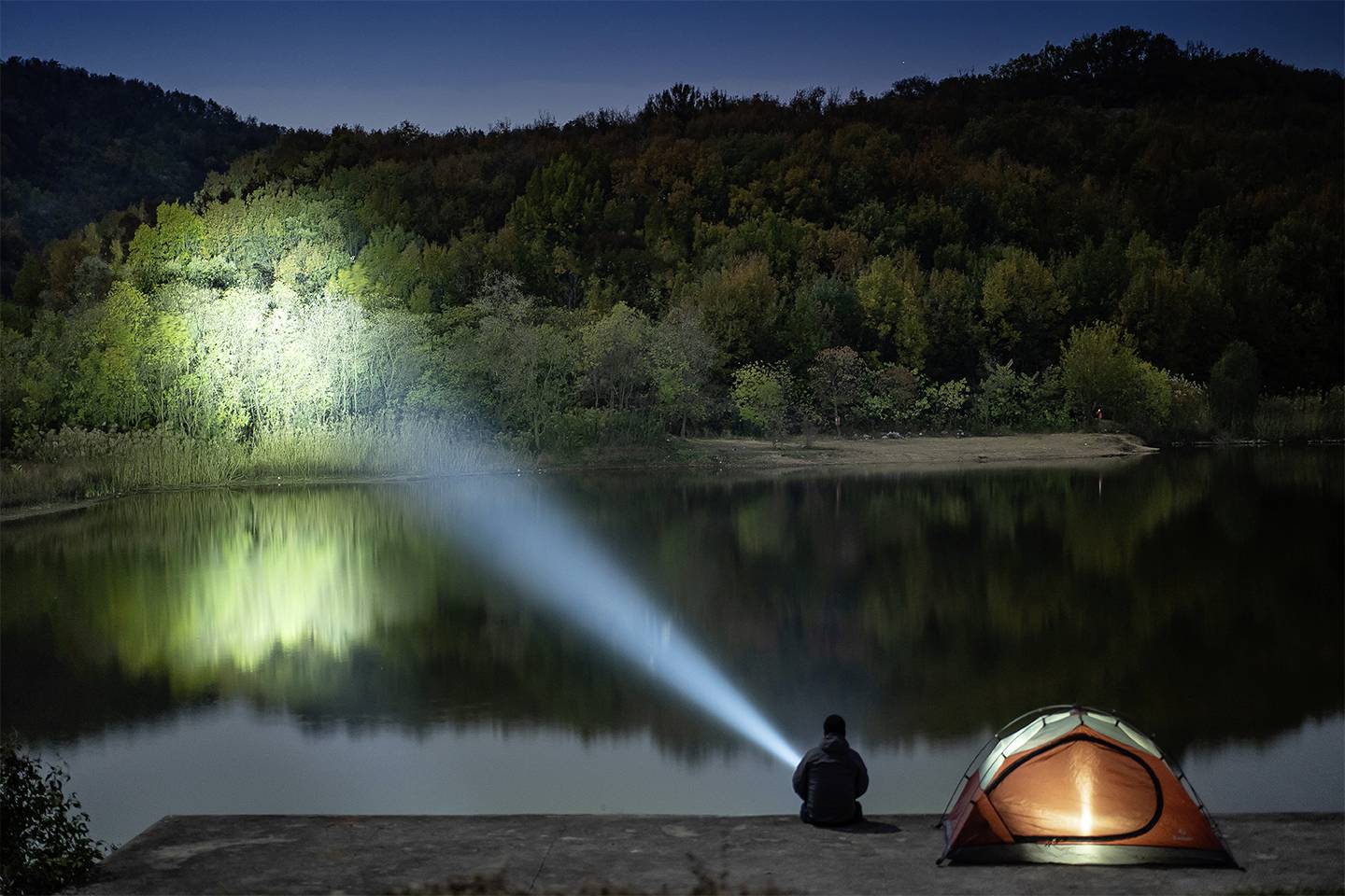 Jedna osoba siedzi nocą nad jeziorem obok oświetlonego namiotu i kieruje latarkę na drzewa, które odbijają się w wodzie.