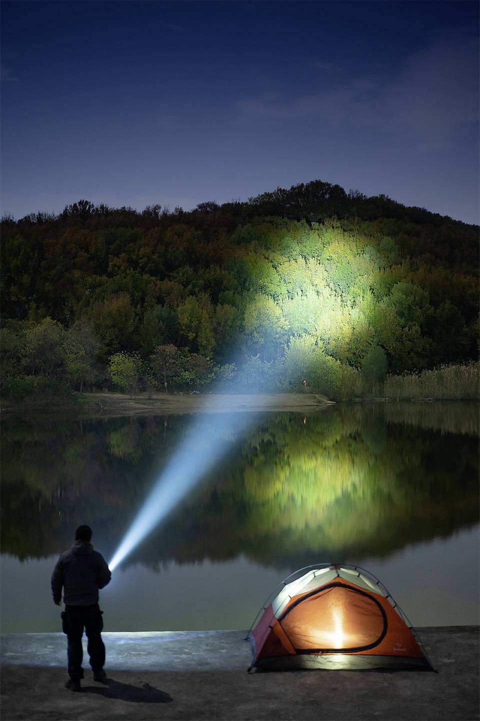 Osoba stoi z latarką nad brzegiem jeziora, oświetlając nocny fragment lasu. Obok stoi namiot z włączonym światłem.
