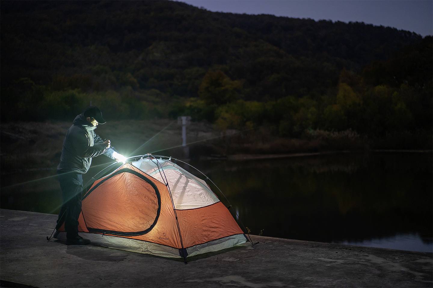 Osoba rozstawia pomarańczowy namiot nocą, używając latarki, nad brzegiem spokojnego jeziora, w otoczeniu zalesionych wzgórz.