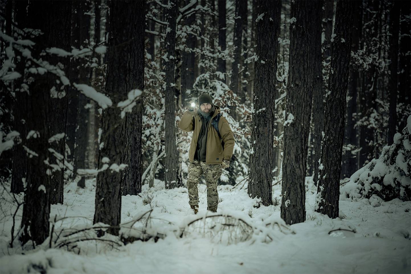Osoba z latarką idzie przez ośnieżony las, otoczona wysokimi drzewami. Scena wygląda na spokojną i odległą od zgiełku.