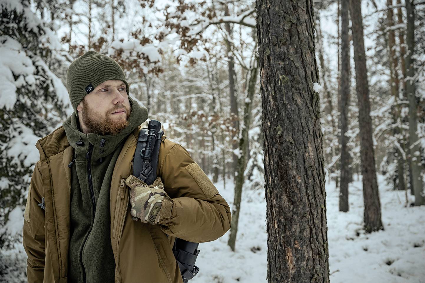 Mężczyzna w stroju zimowym, otoczony ośnieżonym lasem, spogląda w bok. Trzyma czarny plecak i ma na rękach rękawiczki.