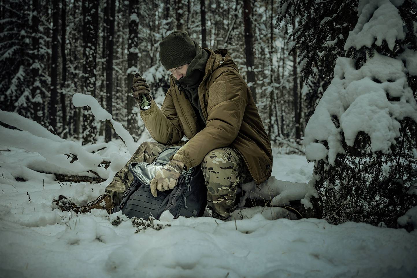 Osoba siedzi w zaśnieżonym lesie, ubrana w ciepłą odzież, otwiera plecak. Otoczenie jest pełne śniegu i drzew iglastych.