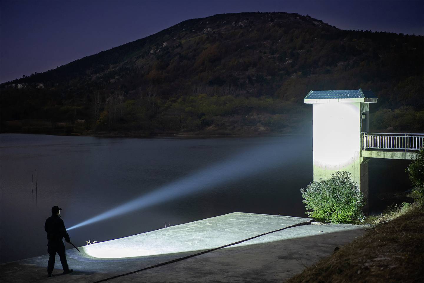 Mężczyzna świeci silną latarką na pomost nad jeziorem w nocy. W tle widać zalesione wzgórze.