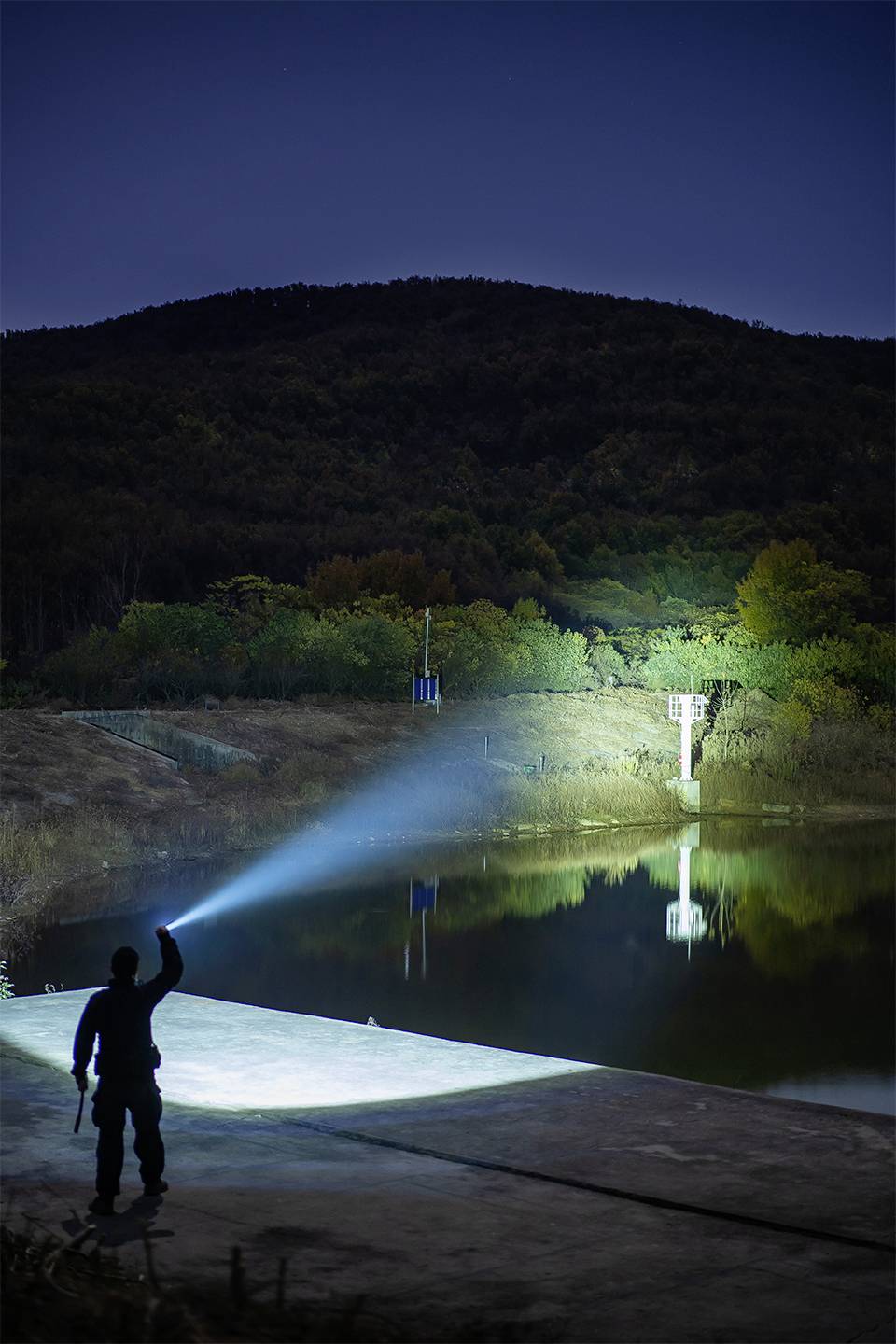 Osoba świeci latarką nocą na jezioro otoczone zalesionymi wzgórzami. Światło odbija się w wodzie.