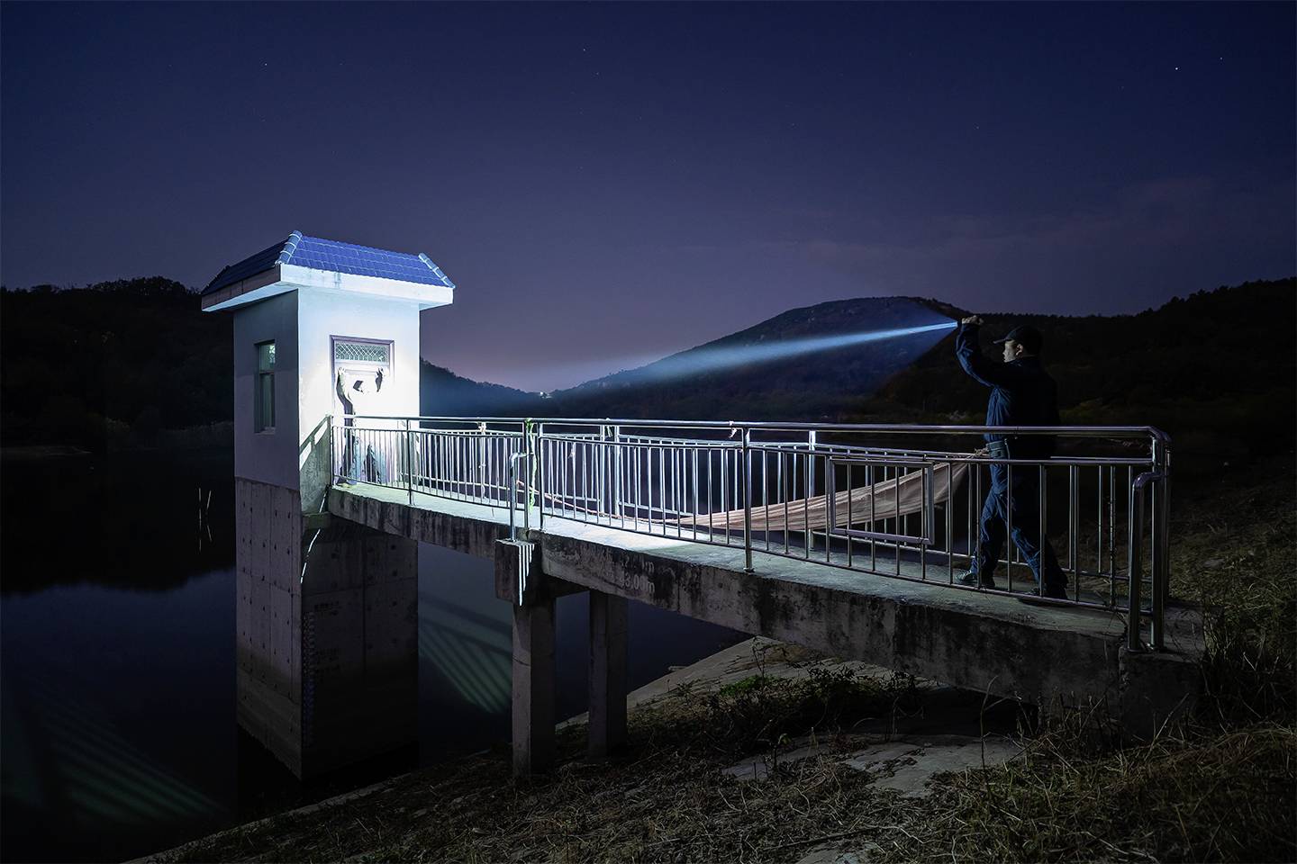Osoba oświetla nocą latarką ścianę budynku na moście nad spokojną taflą wody.
