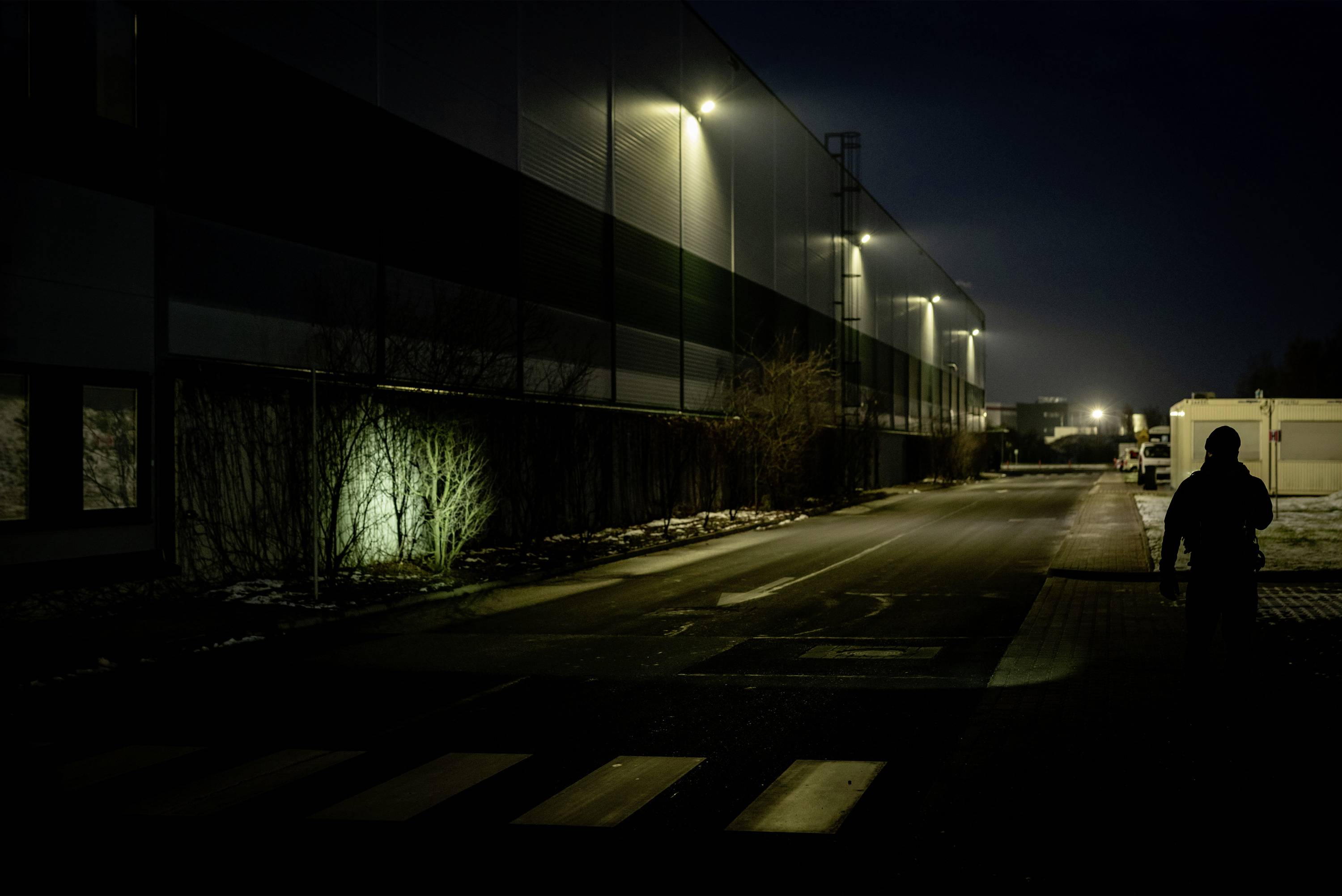Ciemna ulica nocą z oświetlonymi budynkami po lewej stronie. Jedna osoba idzie chodnikiem.